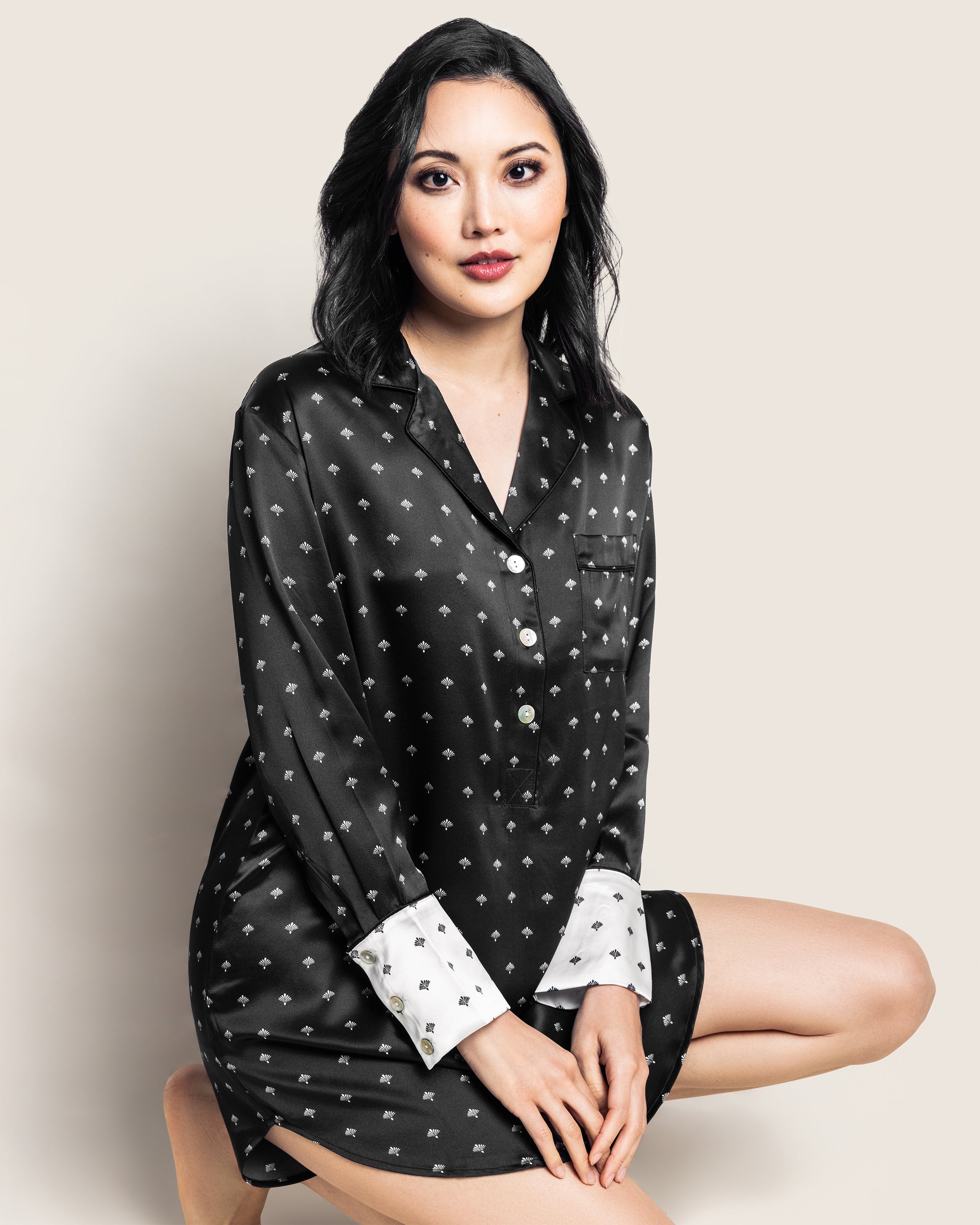A dark-haired woman wears the Womens Silk Nightshirt in Black Art Nouveau by Petite Plume, seated against a neutral background.