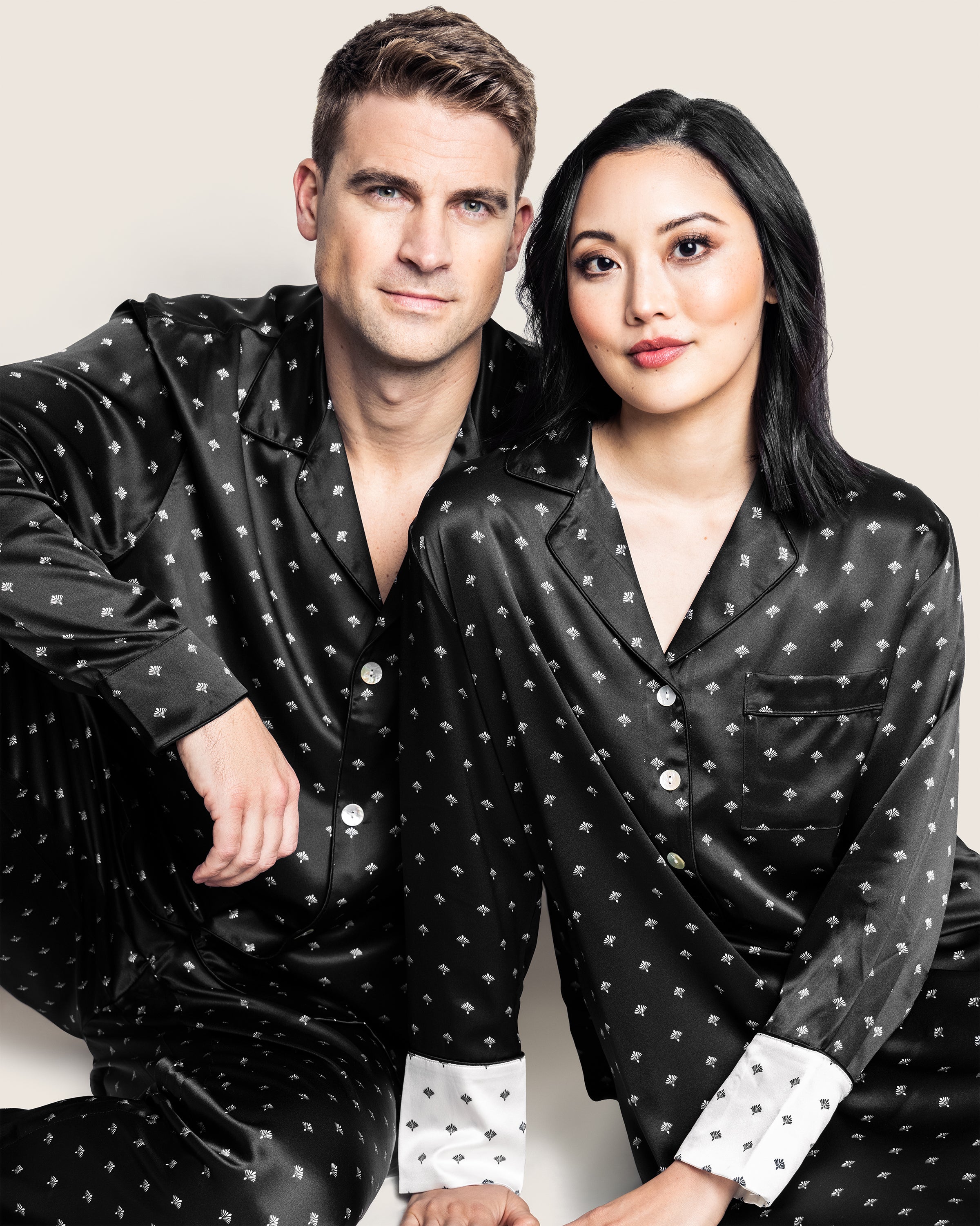 A man and a woman relax together, wearing Petite Plumes matching black pajamas. The womans outfit is the Womens Silk Nightshirt in Black Art Nouveau, featuring a white pattern with crisp white cuffs that highlight their quiet elegance against the plain light background.