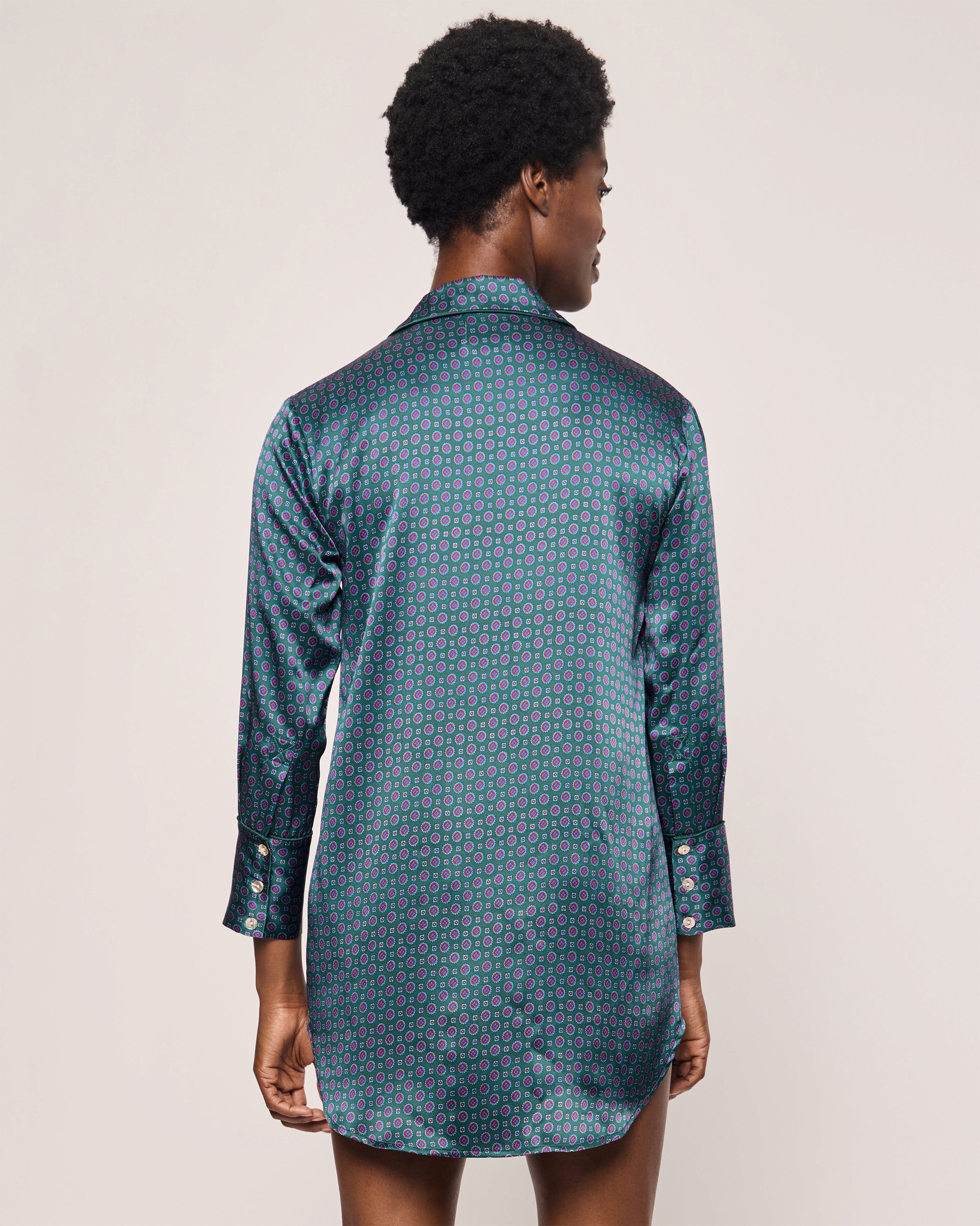 A person with short curly hair stands with their back to the camera wearing Petite Plumes Womens Silk Nightshirt in Emerald Foulard, a long-sleeved dark shirt featuring a blue and pink geometric pattern with buttoned cuffs, complemented by a flowing Mulberry silk neck scarf.