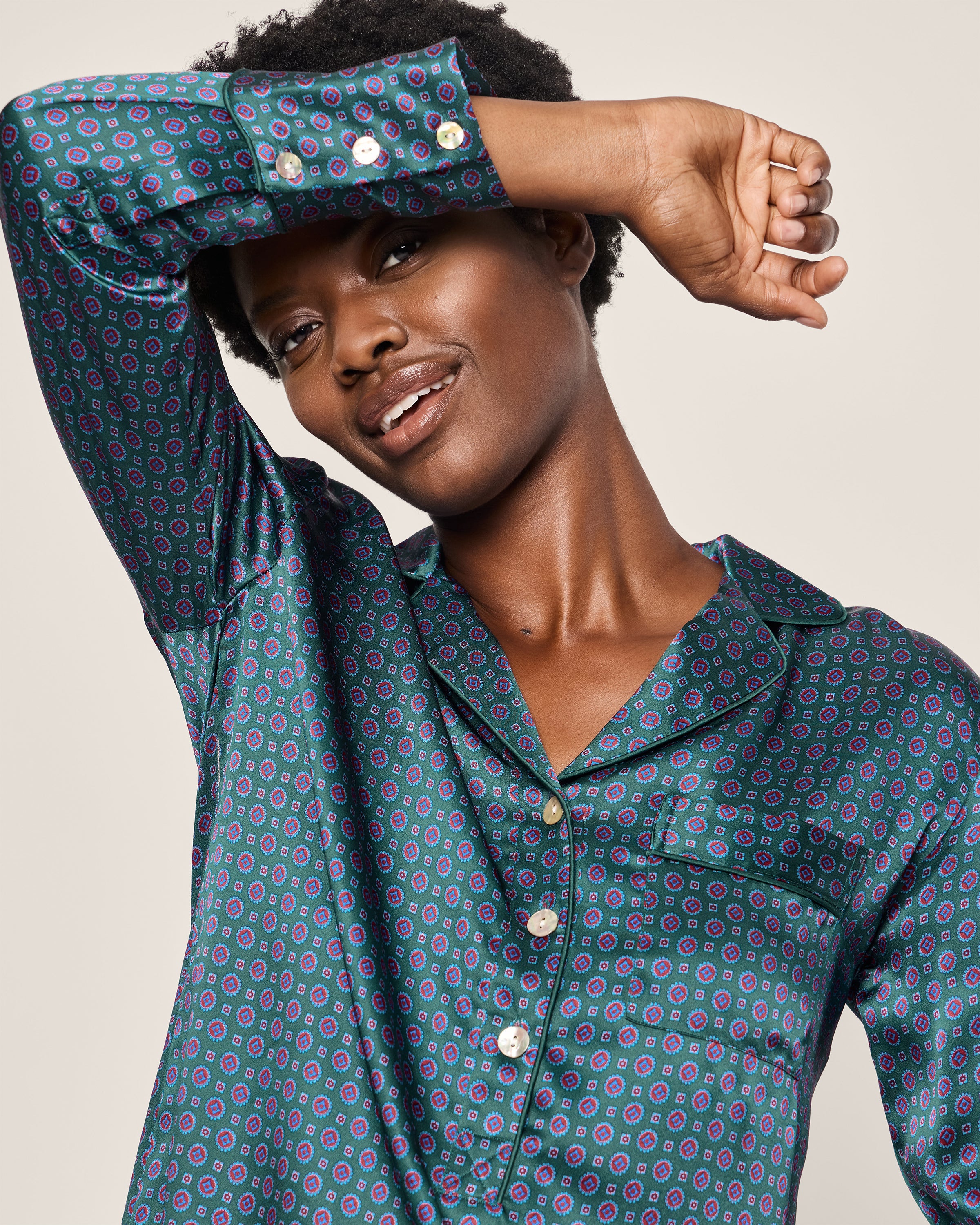 A person with short curly hair smiles, raising one arm above their head. They wear an elegant Womens Silk Nightshirt in Emerald Foulard by Petite Plume, shimmering with a geometric pattern and shiny buttons. A plain, light-colored background highlights the sophistication of their attire.