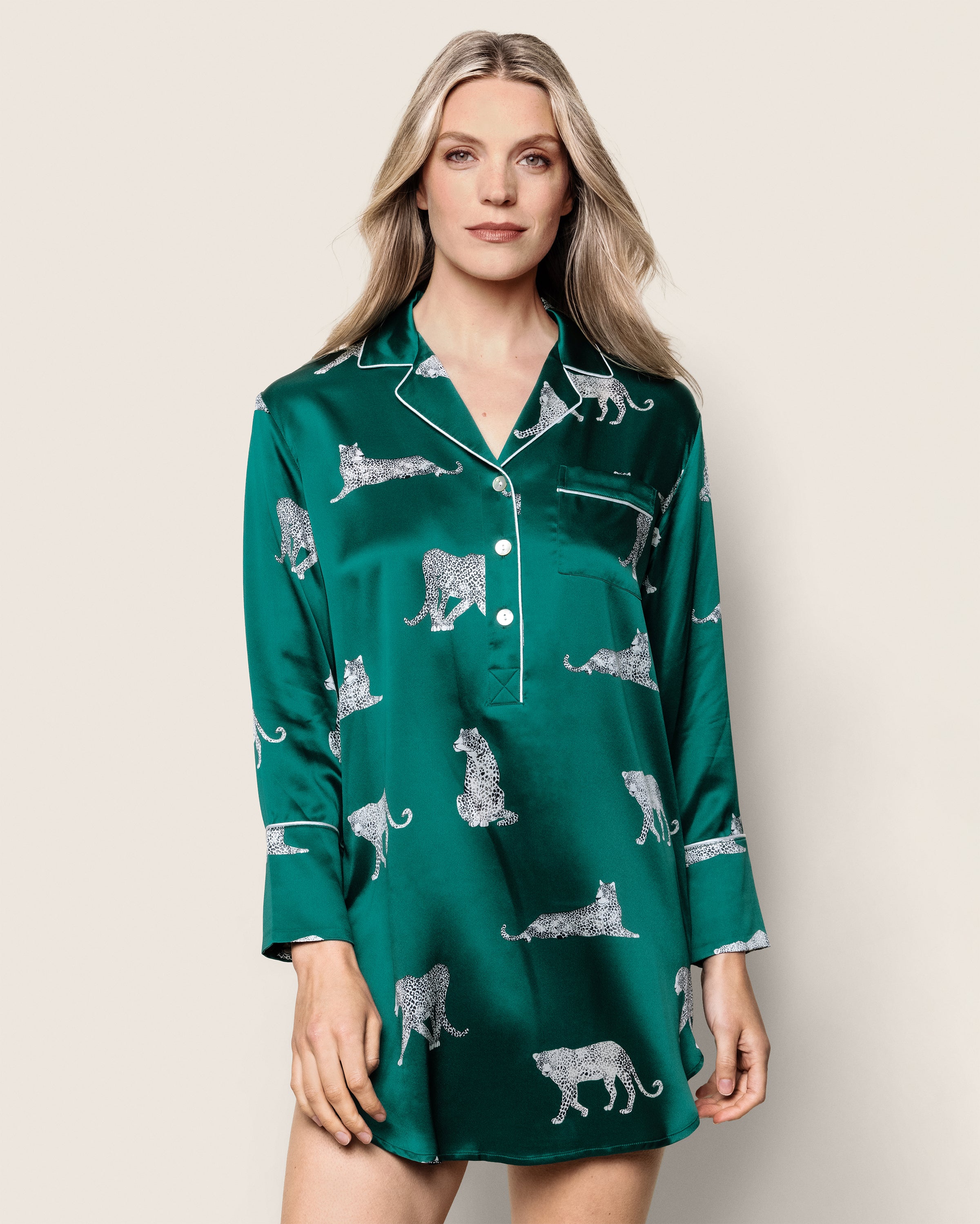 A woman in a Womens Silk Nightshirt in Panthère de Luxe by Petite Plume exudes elegance. The mulberry silk features green with leopards, white piping, and buttons, set against a plain background. Her loose waves perfectly complement the luxurious sleepwear ensemble.