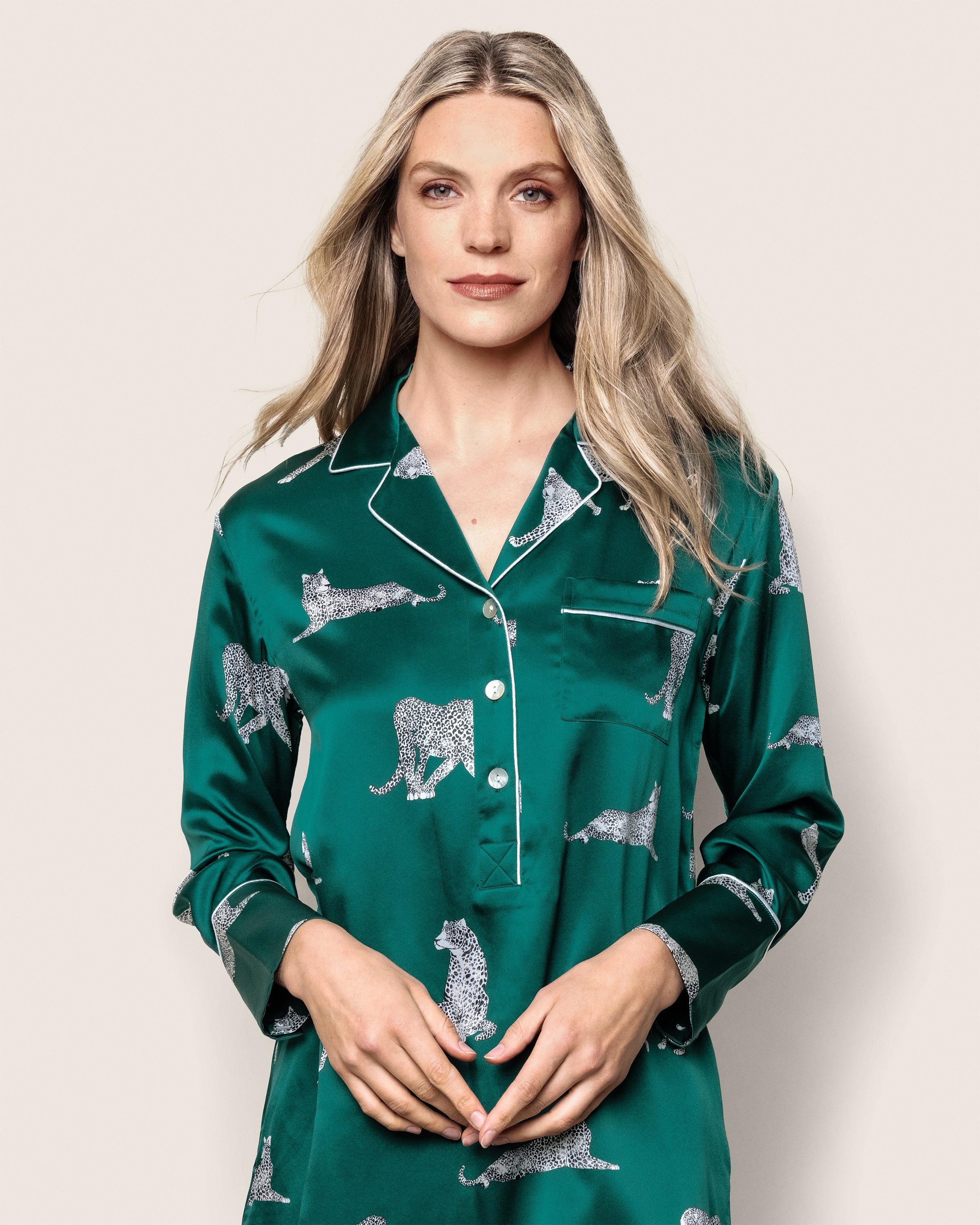 A woman with long blonde hair wears a luxurious Womens Silk Nightshirt in Panthère de Luxe by Petite Plume, crafted from mulberry silk and adorned with white animal motifs, standing elegantly against a plain light background.