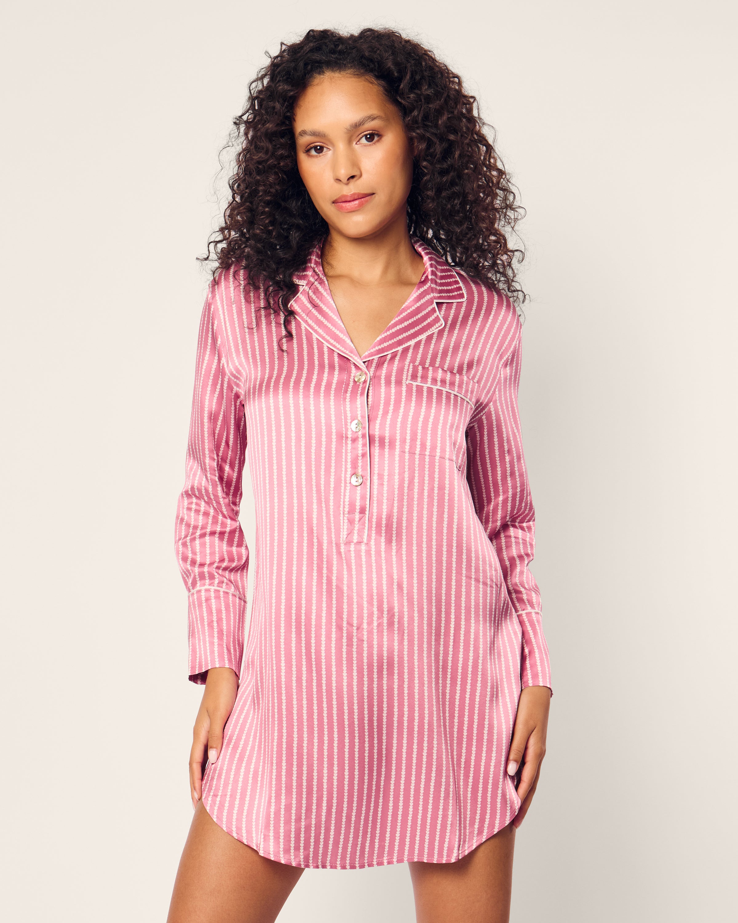 A woman with curly hair models the exquisite Womens Silk Nightshirt in Love Lines by Petite Plume. The tailored pink striped satin shirt dress features a collar and button placket, as she confidently gazes at the camera against a neutral background.