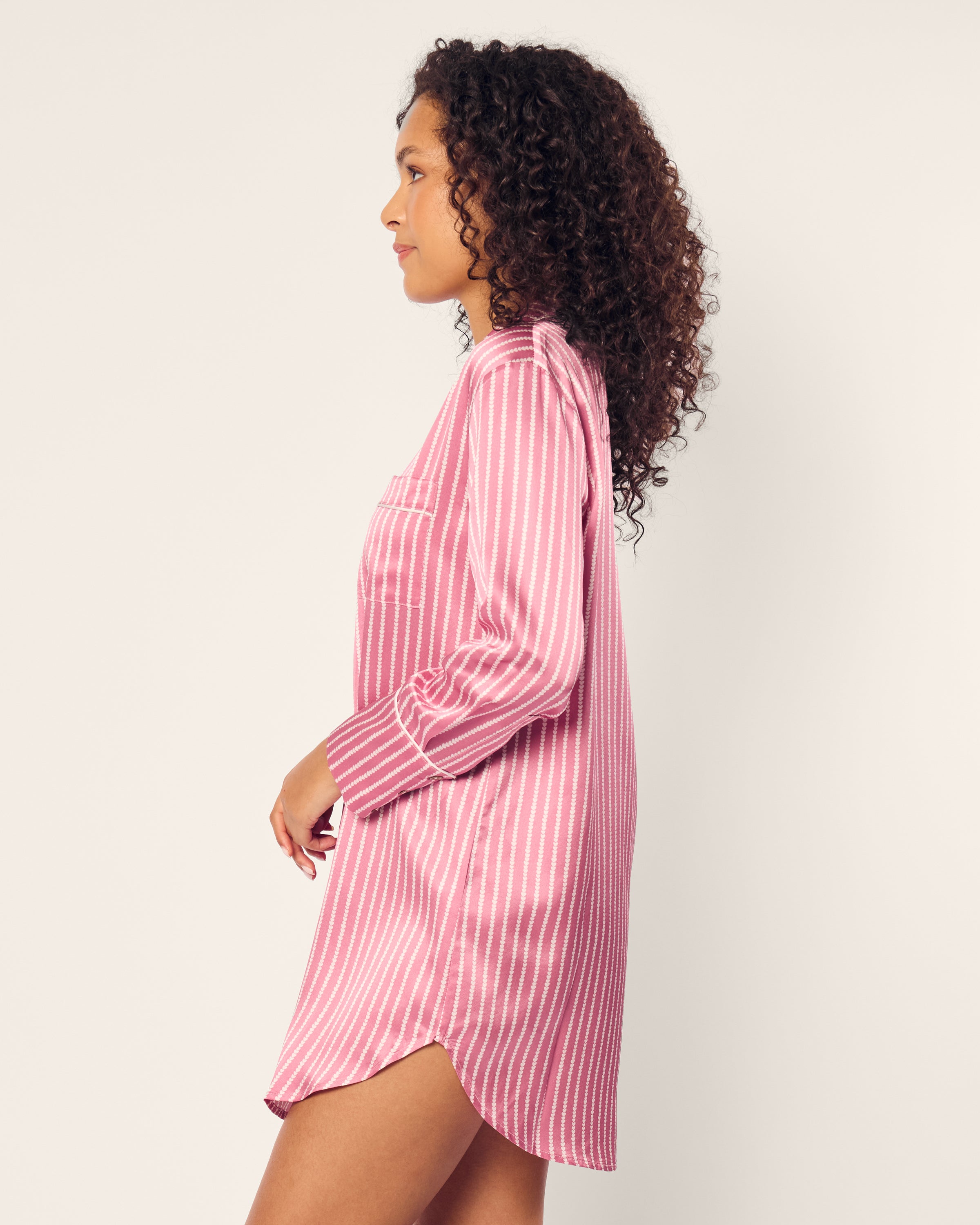 A person with curly hair wears the Womens Silk Nightshirt in Love Lines by Petite Plume, showcasing pink and white vertical stripes, while standing sideways against a plain background.