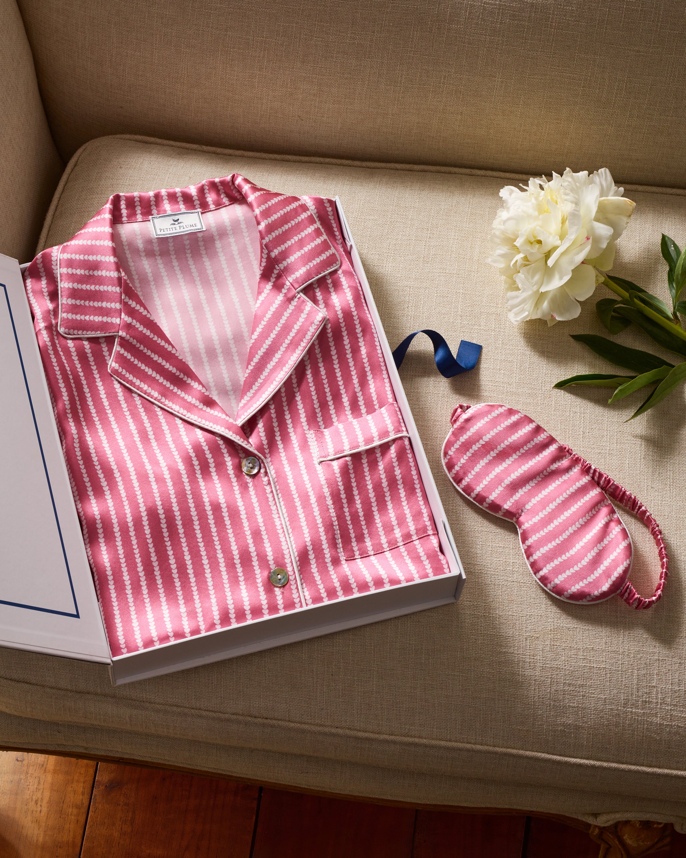 A beige sofa holds an open box with Petite Plumes Womens Silk Nightshirt in Love Lines, accompanied by a matching mulberry silk sleep mask and a white peony with green leaves.