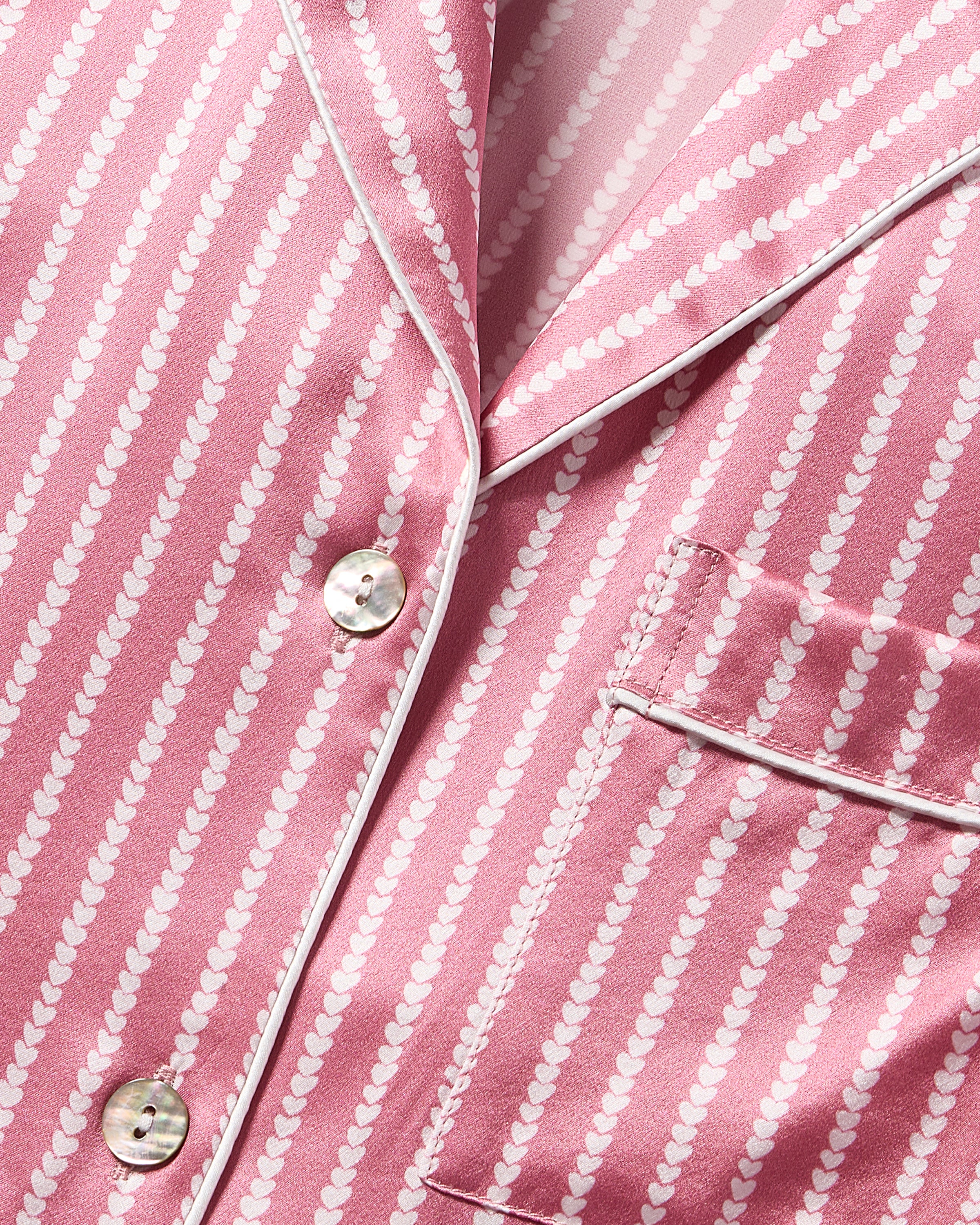 Close-up of the Womens Silk Nightshirt in Love Lines by Petite Plume, crafted from luxurious mulberry silk with pink fabric and white vertical dotted stripes. This tailored design showcases a silky texture, featuring a chest pocket and white buttons, with elegant piping on the collar and pocket edge.