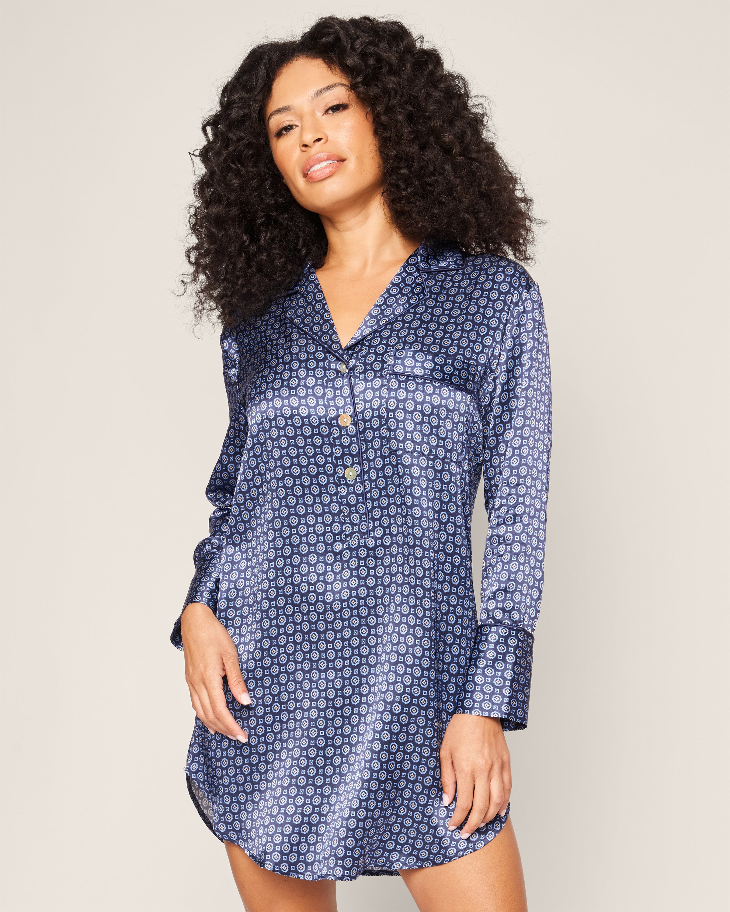 A person with long, curly hair is wearing Petite Plumes Womens Silk Nightshirt in Midnight Foulard. The shirt dress features a stylish button-down design, and they stand against a plain light background, gazing confidently at the camera.