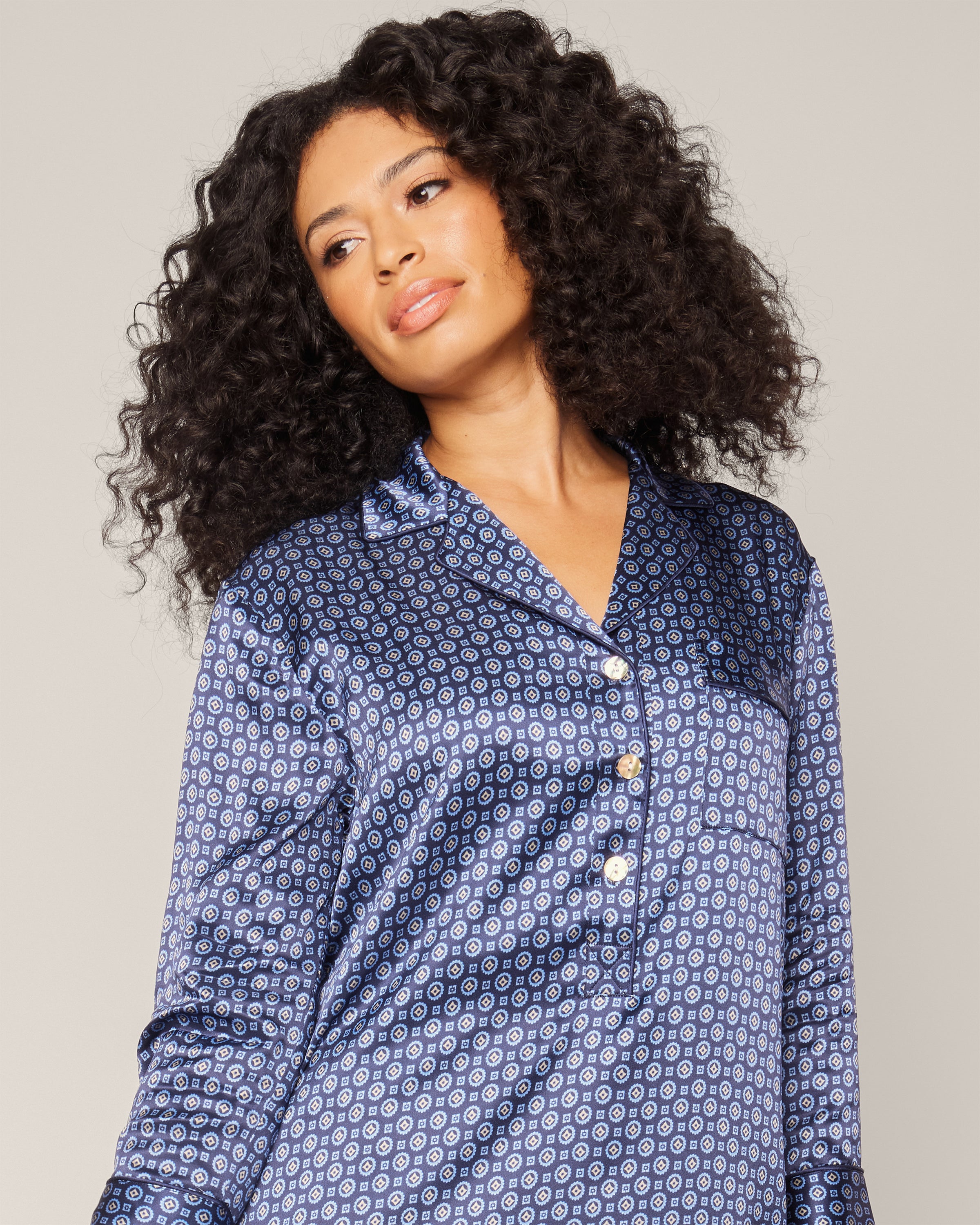 A person with long, curly hair wearing a Petite Plume Womens Silk Nightshirt in Midnight Foulard and a stylish neck scarf stands against a plain background, looking slightly to the side.