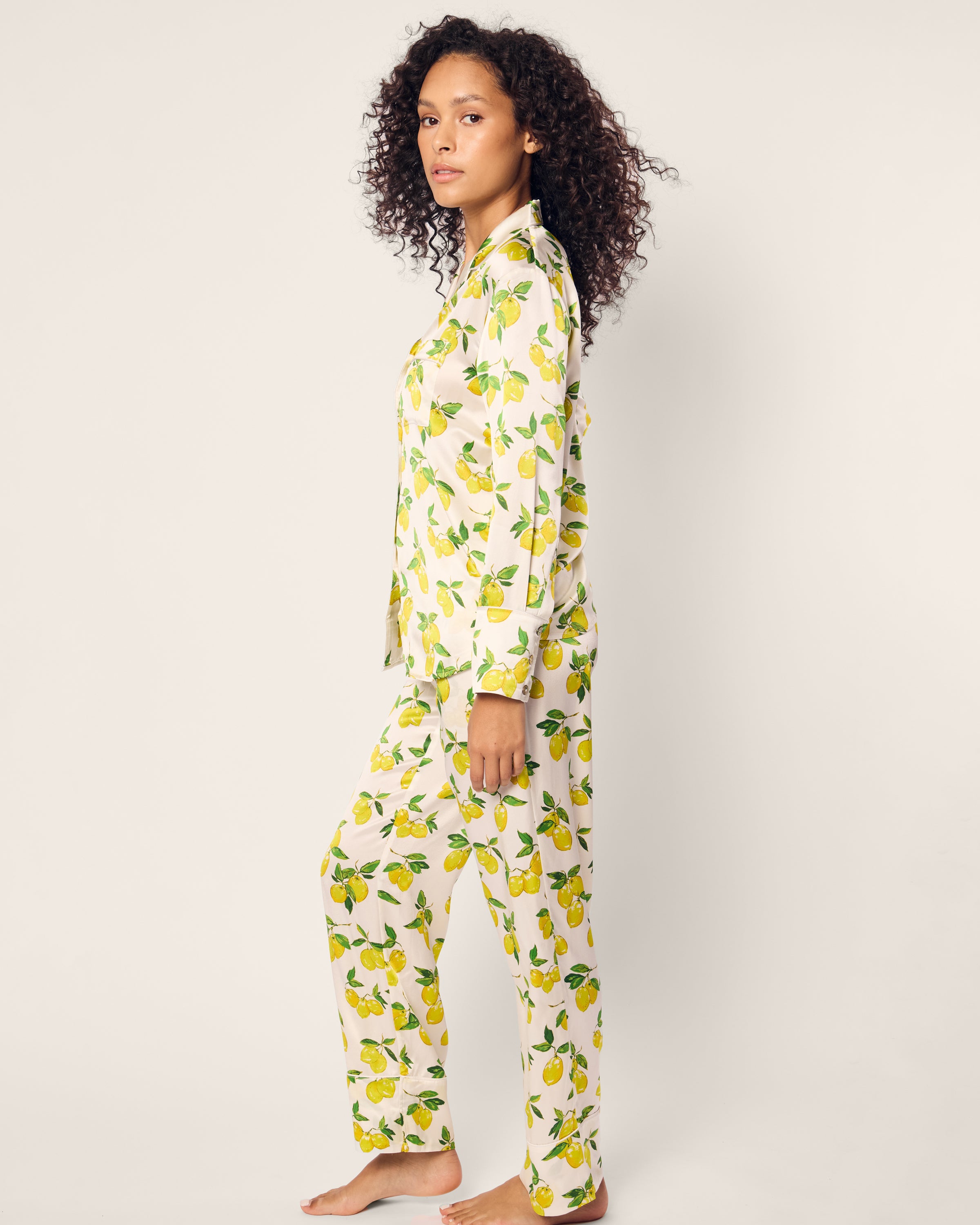 A curly-haired person stands barefoot wearing Petite Plumes Womens Silk Pajama Set in Amalfi Lemons. The light-colored outfit with yellow lemons and green leaves contrasts against the plain, neutral background.