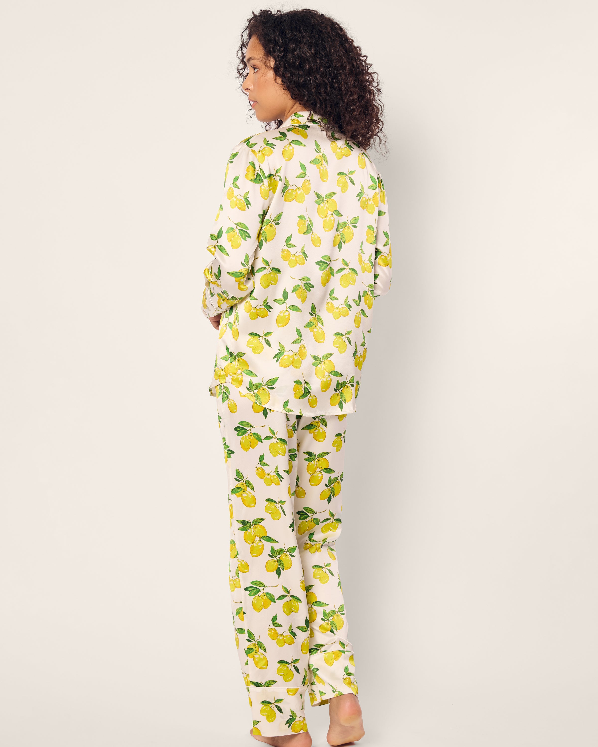 Curly-haired and barefoot, a person wears the Petite Plume Womens Silk Pajama Set in Amalfi Lemons, featuring a lemon pattern on a light background. They glance over their shoulder with a neutral expression, embodying an elegant mix of comfort and style.