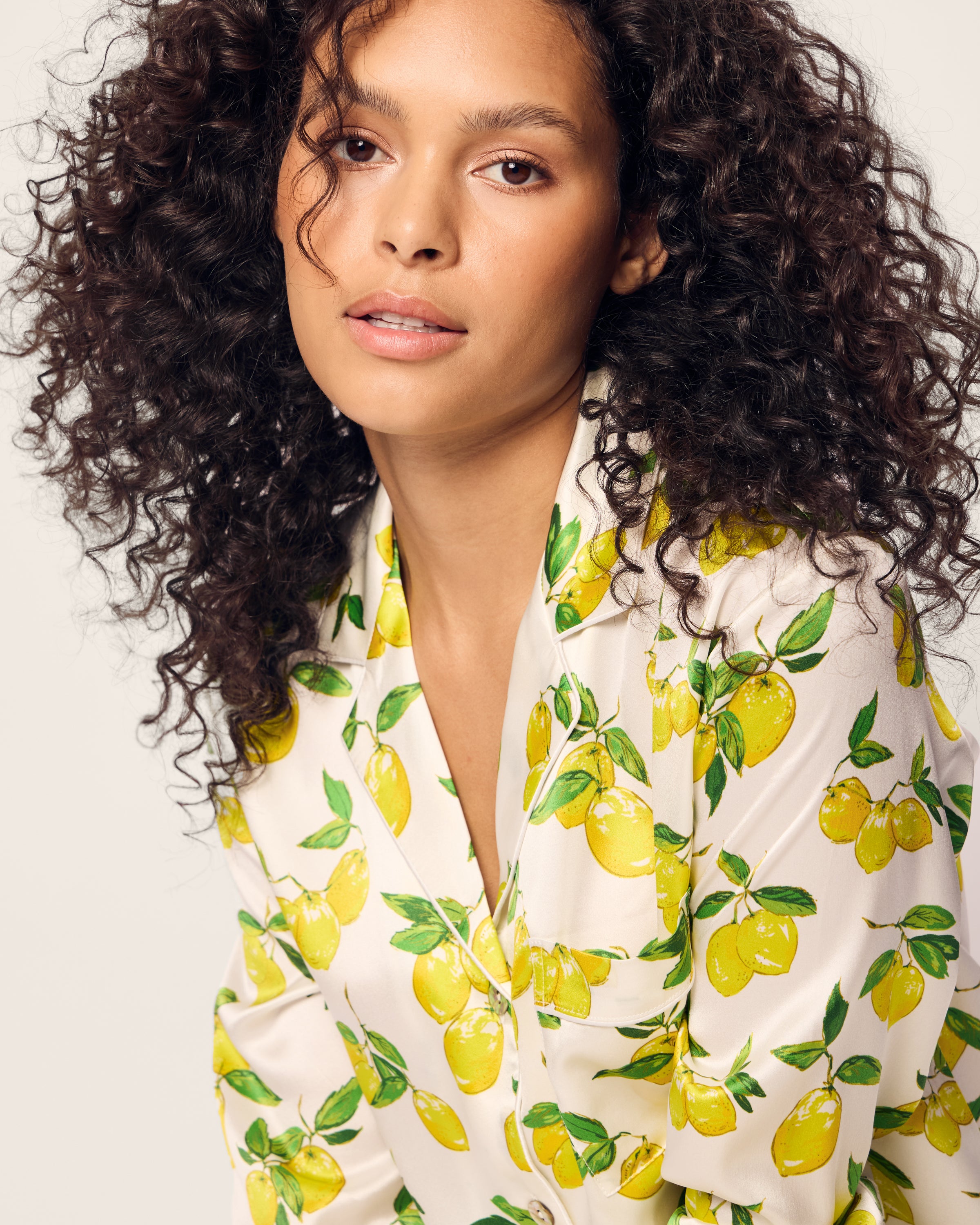 A person with curly hair wearing the limited-edition Petite Plume Womens Silk Pajama Set in Amalfi Lemons looks directly at the camera. The soft, neutral background enhances the luxurious feel of the machine-washable silk fabric.