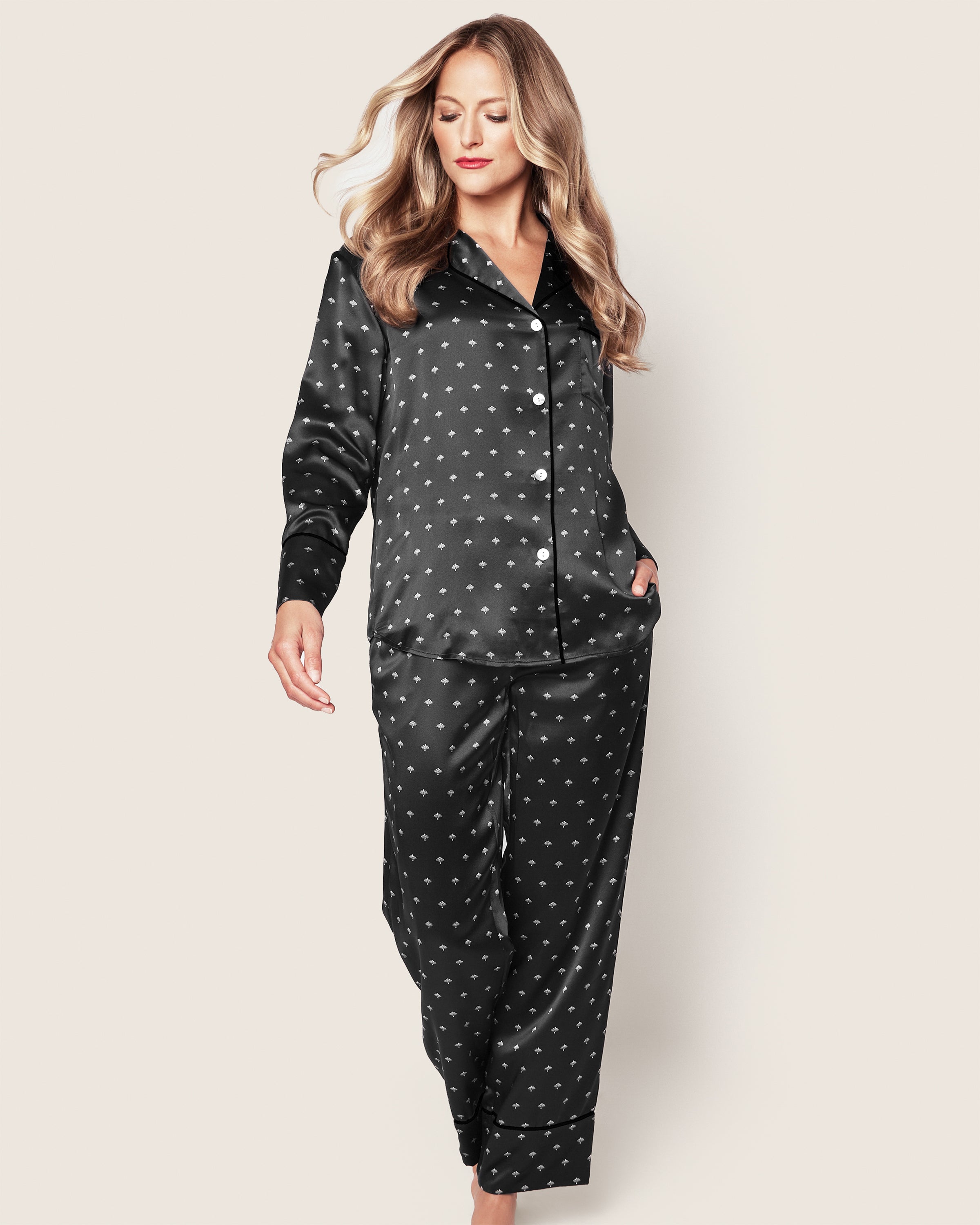 A woman with long, wavy blonde hair models the Petite Plume Womens Silk Pajama Set in Art Nouveau Black, featuring a small white dotted pattern, hands in pockets, against a plain light background.