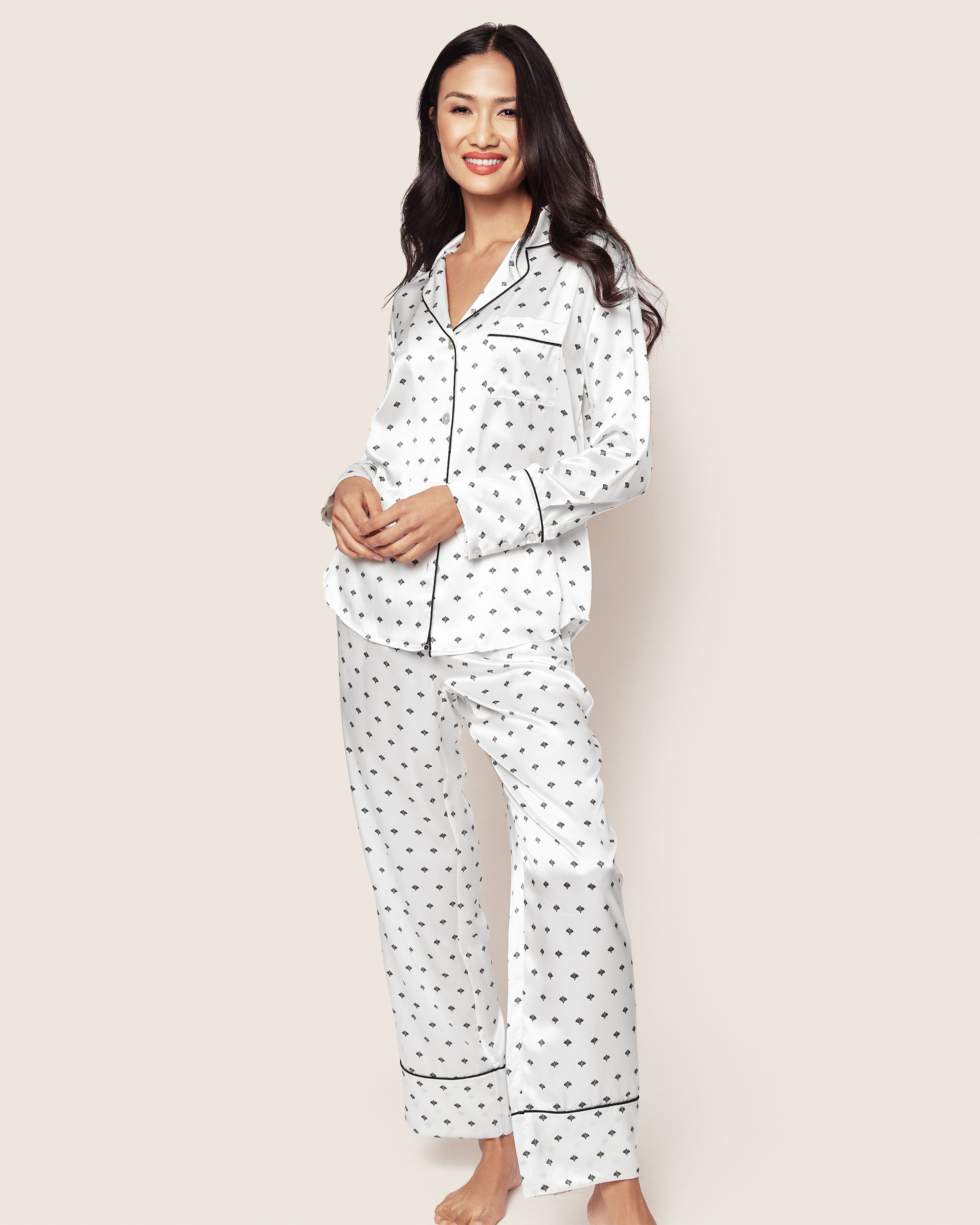 A smiling woman poses in a luxurious Womens Silk Pajama Set from Petite Plume, featuring a white art nouveau design. Her long, dark hair contrasts elegantly with the sets button-up top and pants, both trimmed with black piping, against a plain off-white backdrop.