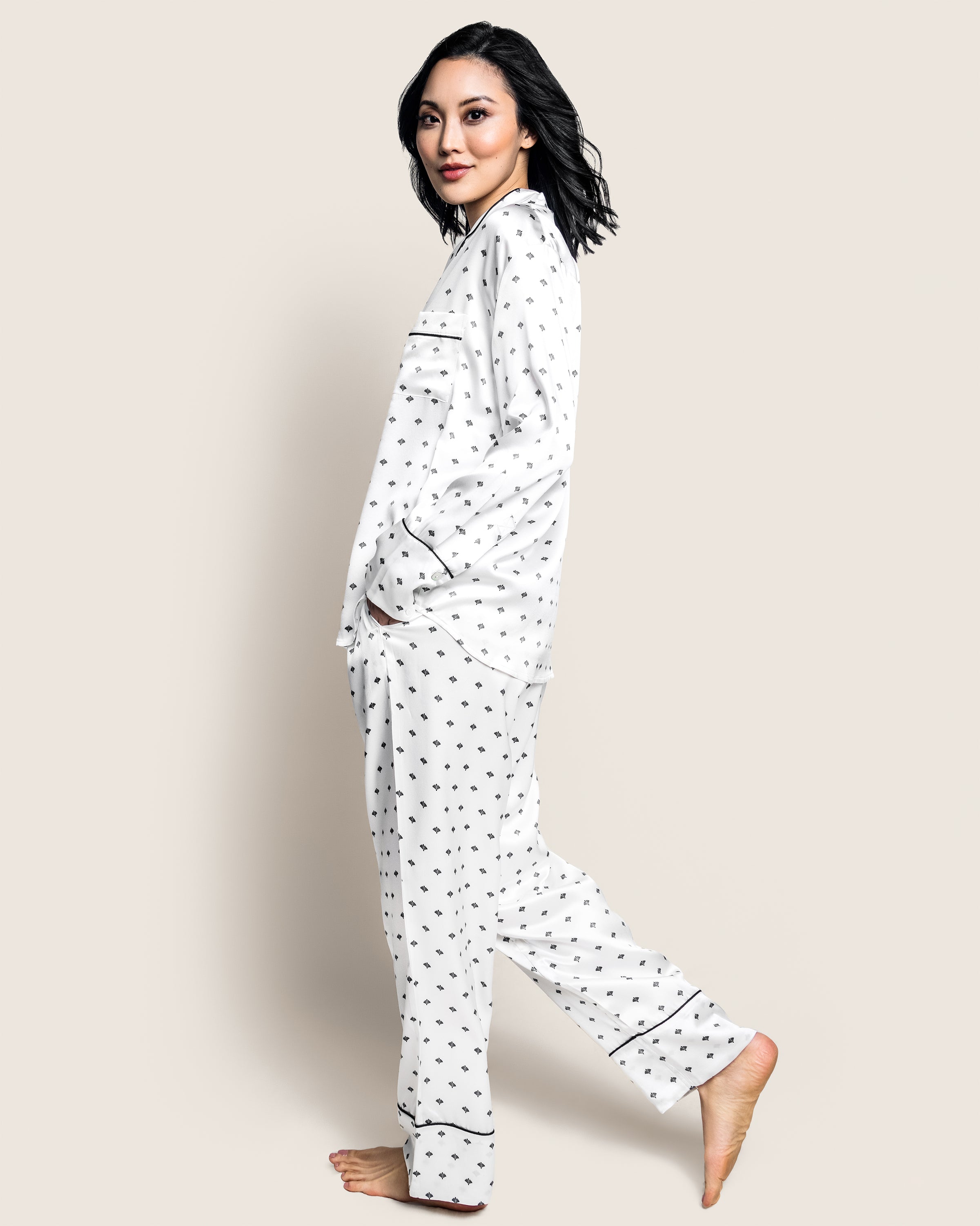 A person with dark hair stands barefoot against a light background, wearing the Womens Silk Pajama Set in White Art Nouveau by Petite Plume. They smile at the camera, their left hand in a pocket, exuding an old Hollywood charm.