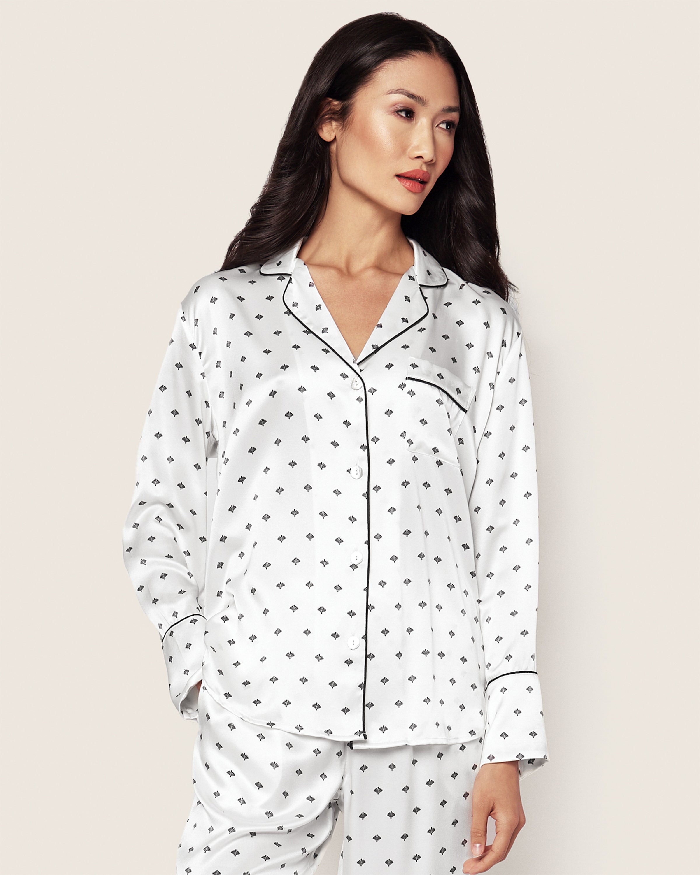 A woman models a Petite Plume Womens Silk Pajama Set in White Art Nouveau with black piping, standing against a plain backdrop. With long dark hair and an expressionless face, her style evokes old Hollywood glamour.