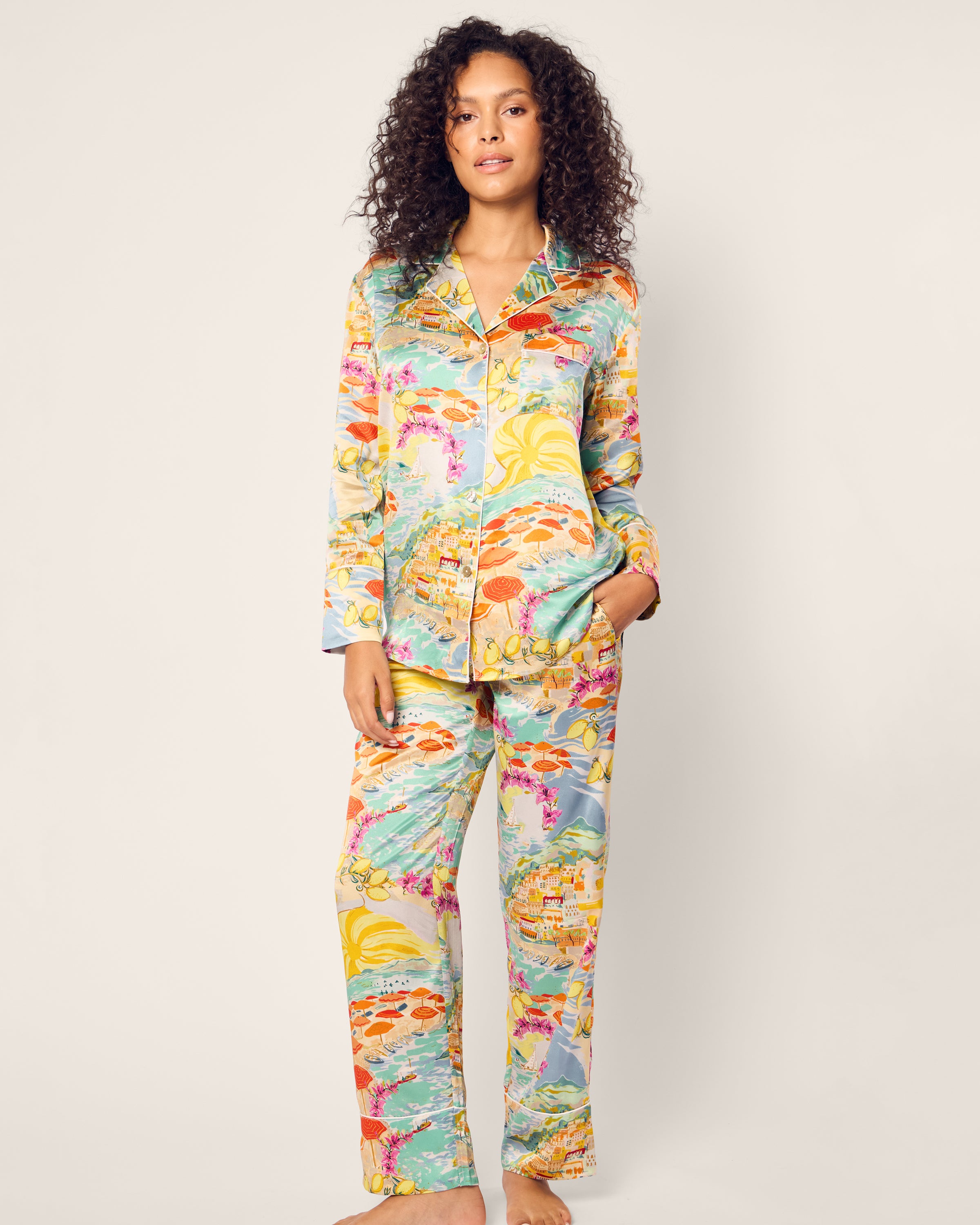 A person in the luxurious Womens Silk Pajama Set in La Dolce Vita by Petite Plume stands against a plain background. Their curly hair frames their face as they gaze forward, with one hand casually resting in their pocket.