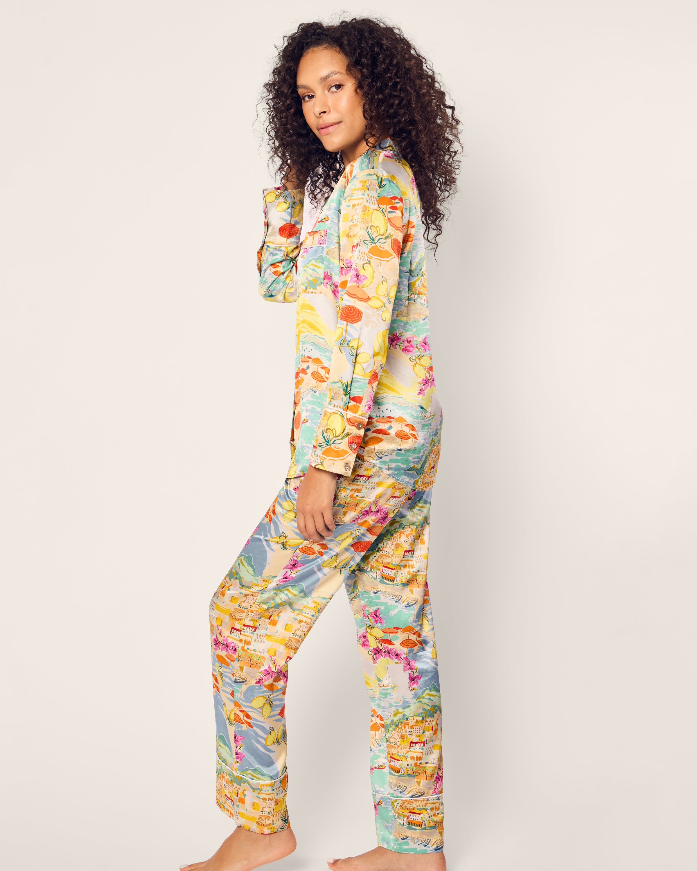 A person with curly hair stands barefoot in Petite Plumes colorful Womens Silk Pajama Set in La Dolce Vita, featuring a floral and tropical theme. They face sideways with a relaxed expression, one hand gently touching their hair, against a plain off-white background.