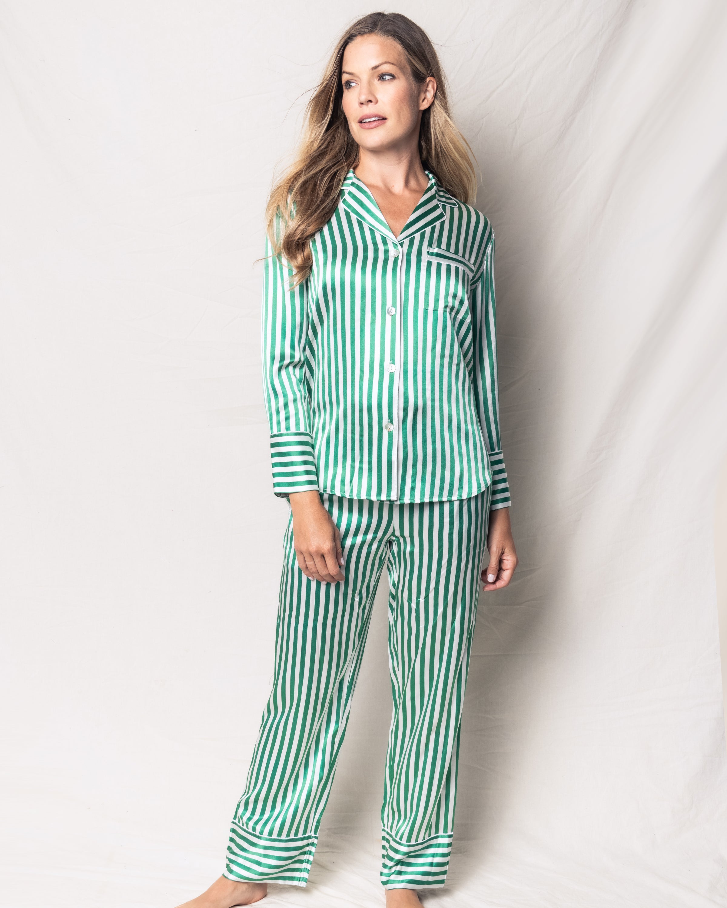 Dressed in Petite Plumes luxurious Womens Silk Pajama Set in Green Stripe, a person stands against a light background. The mulberry silk pajamas feature long sleeves, a collar, and full-length pants. Their hair is styled loosely as they gaze slightly to the side.
