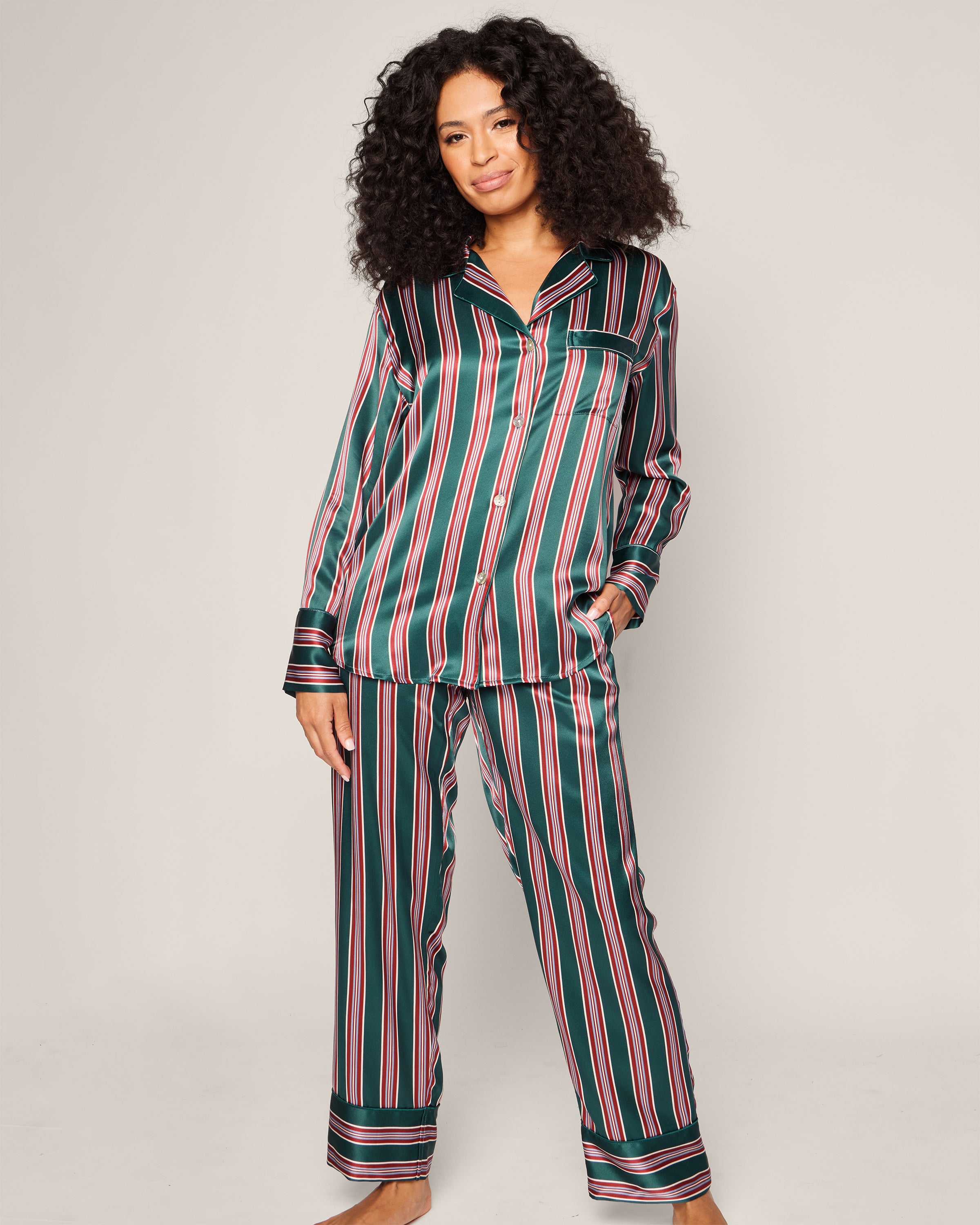 A woman with curly hair wears the luxurious Petite Plume Womens Silk Pajama Set in Lancaster Stripe. The hypoallergenic satin-finish set features vertical black, pink, and green stripes with a collared button-down shirt. She stands barefoot against a neutral background.