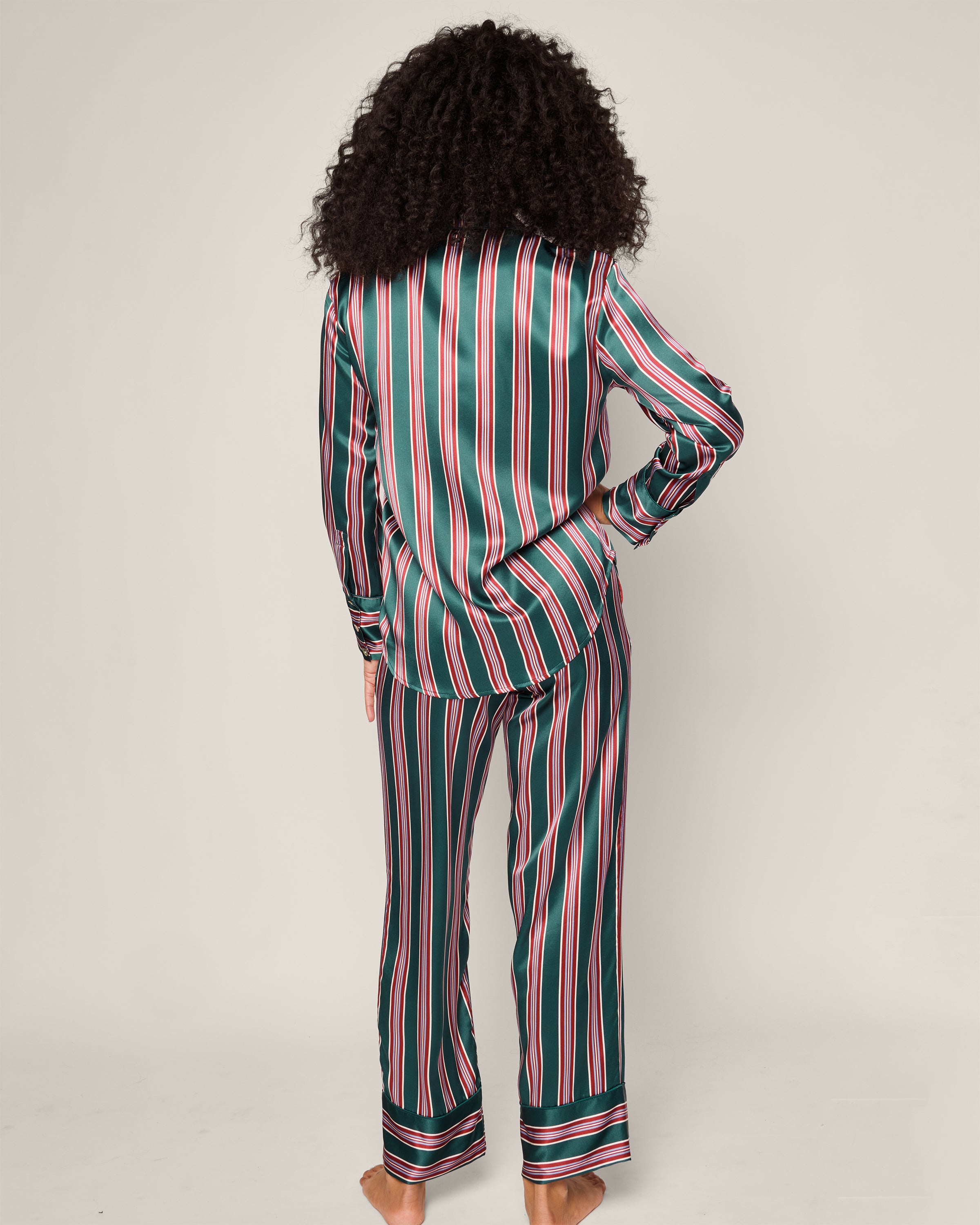 A person with curly hair stands barefoot facing away, wearing Petite Plumes Womens Silk Pajama Set in Lancaster Stripe. This luxurious sleepwear features green, black, pink, and white vertical stripes and is crafted from exquisite hypoallergenic mulberry silk for ultimate comfort and elegance.