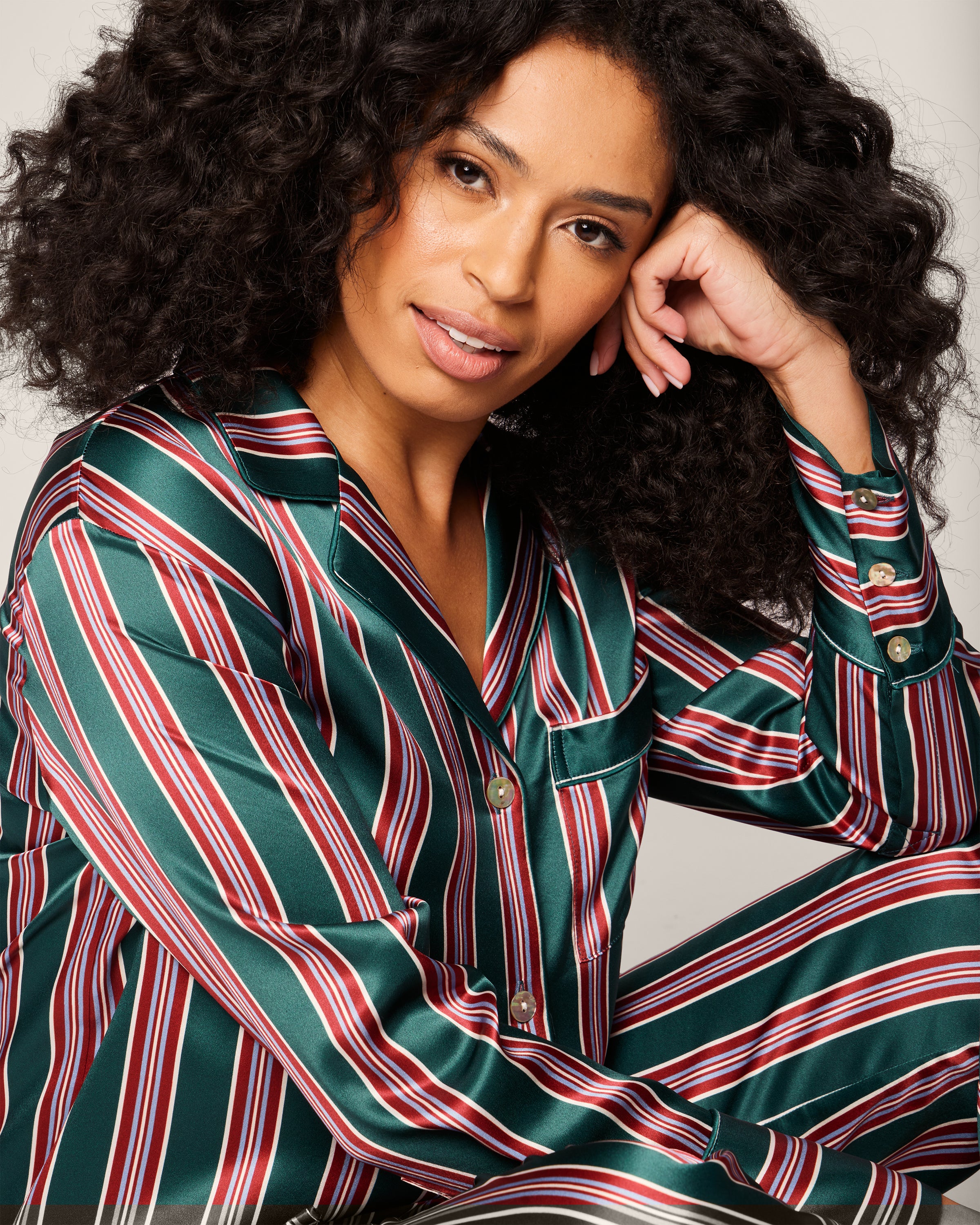 A curly-haired person sits wearing a luxurious Womens Silk Pajama Set in Lancaster Stripe by Petite Plume, adorned with green, white, and red stripes. They smile gently, resting their head on one hand.