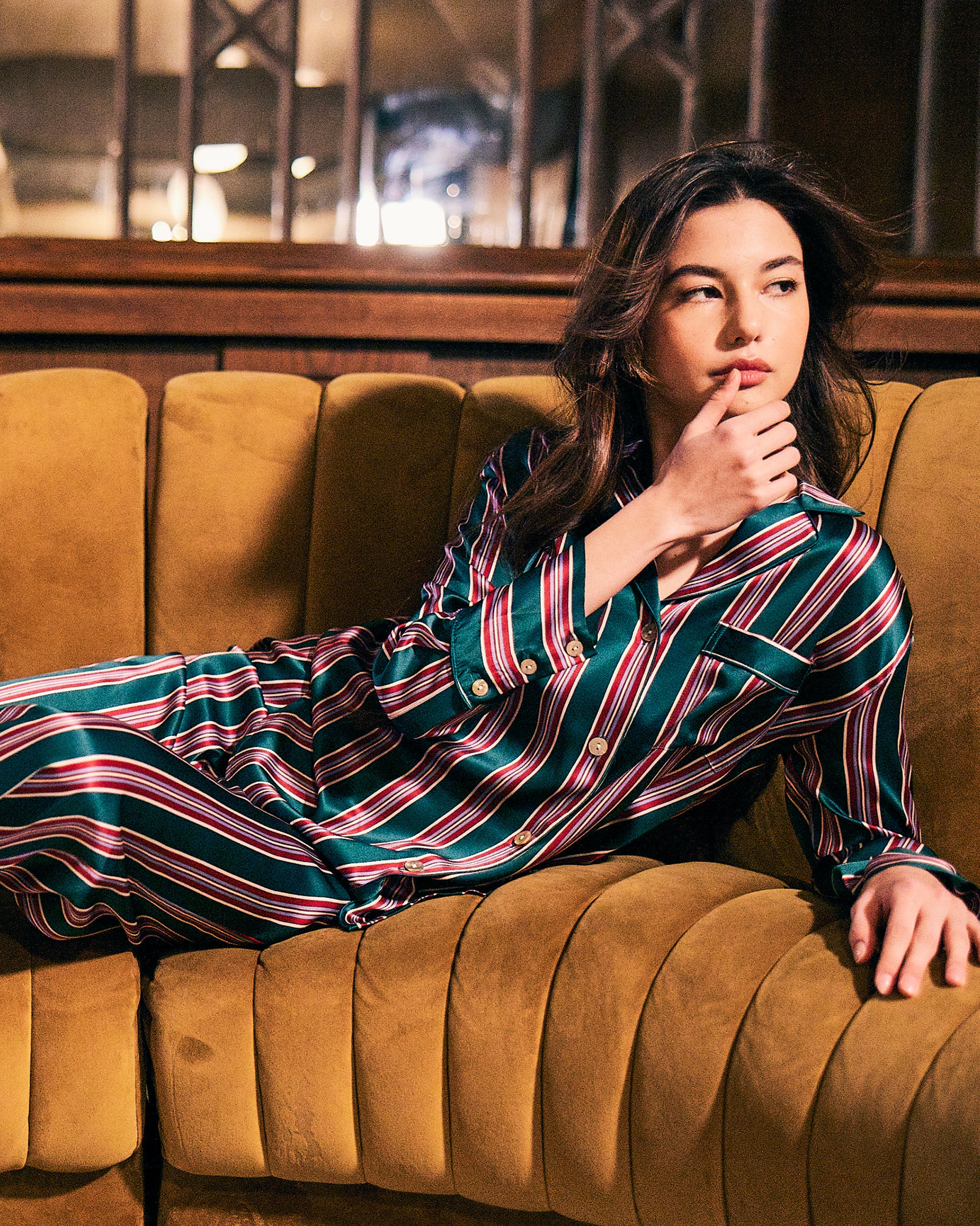Lounging on a plush mustard couch, a person is in luxury sleepwear from Petite Plume—a Womens Silk Pajama Set in Lancaster Stripe. Their dark hair cascades as they rest their chin on one hand, gazing thoughtfully to the side in warm, cozy lighting.
