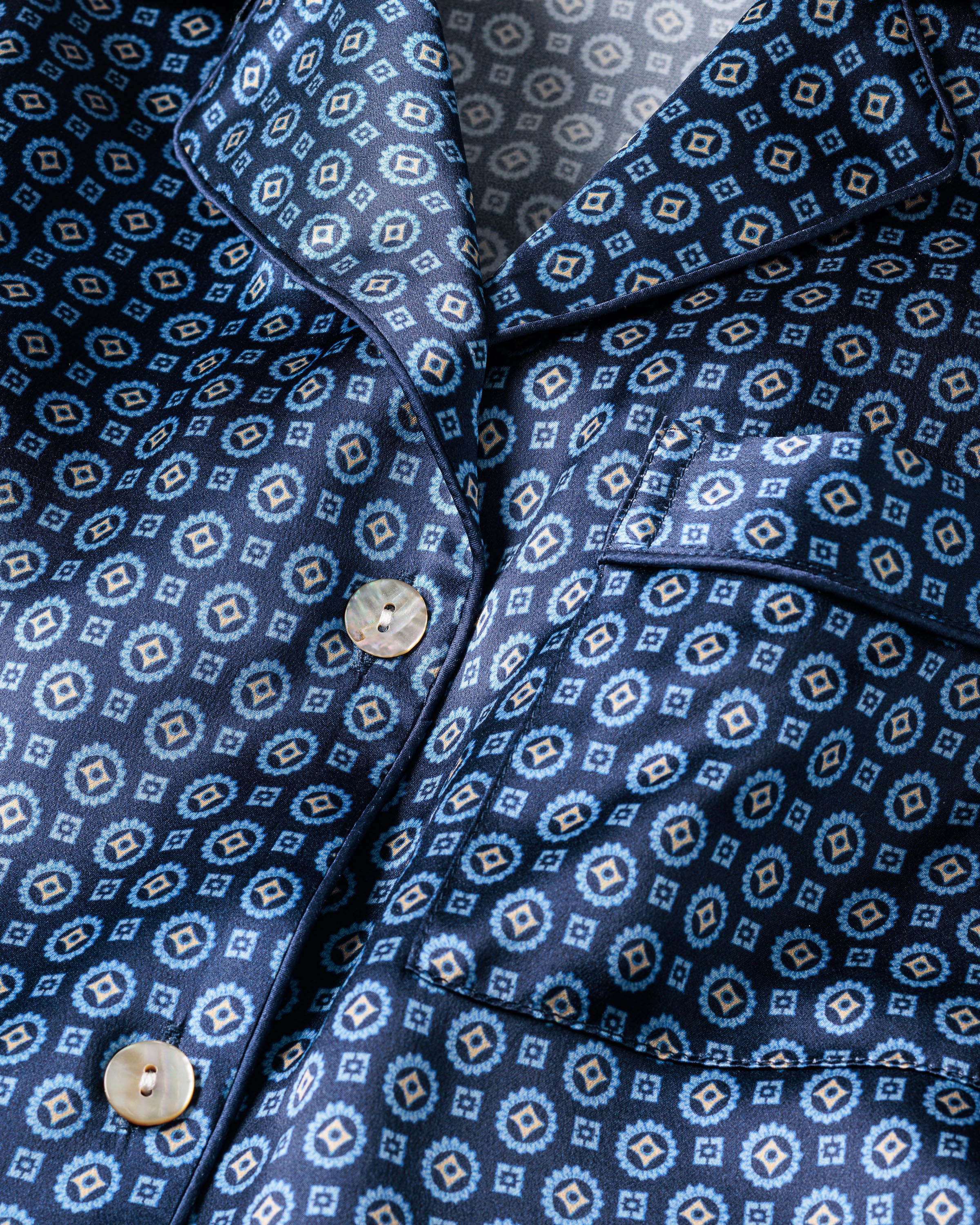 Close-up of the Womens Silk Pajama Set in Midnight Foulard by Petite Plume, showcasing a silky blue geometric pattern with diamond shapes. This luxurious sleepwear features a pocket and cream-colored buttons, with light accentuating its shiny, temperature-regulating Mulberry silk fabric.