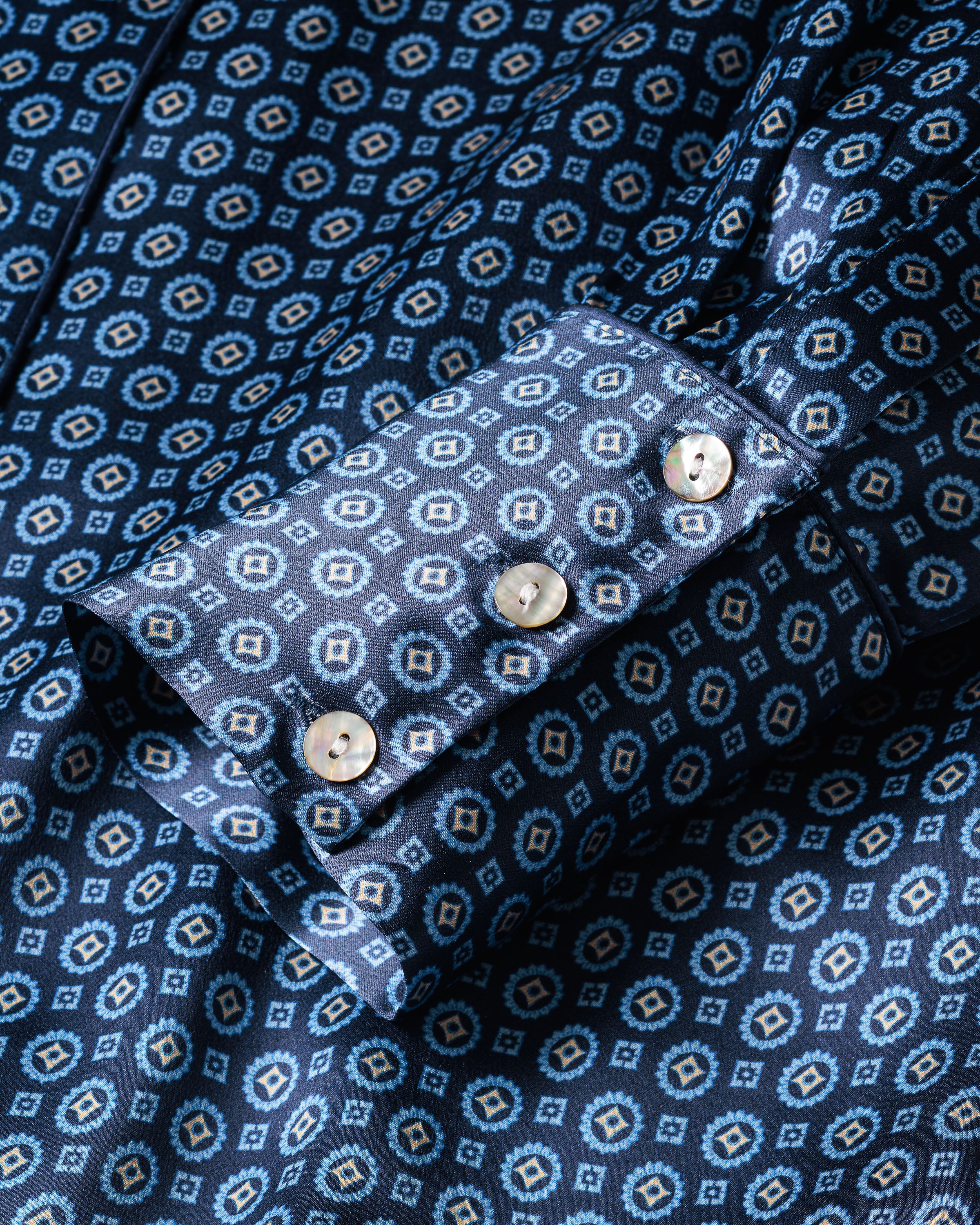 Close-up of the Womens Silk Pajama Set in Midnight Foulard by Petite Plume, showcasing a blue fabric with a geometric pattern in lighter blue and beige. The luxurious sleeve is adorned with a cuff featuring four elegant beige buttons, enhancing its sophisticated design.