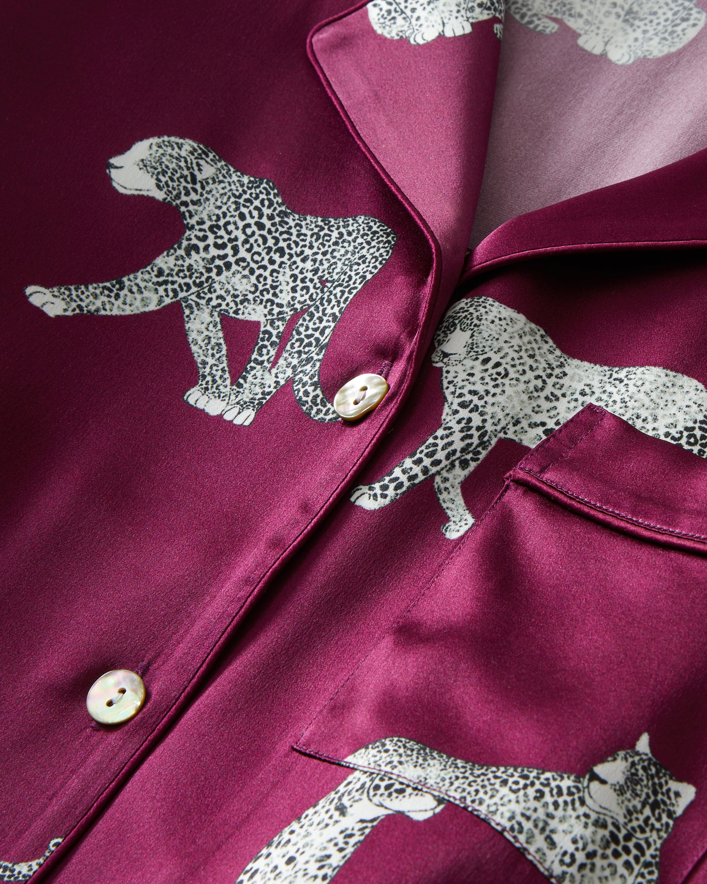 The Women's Silk Pajama Set in Merlot Panthère features a silky, deep magenta button-up shirt and pants adorned with an all-over white illustrated leopard print.