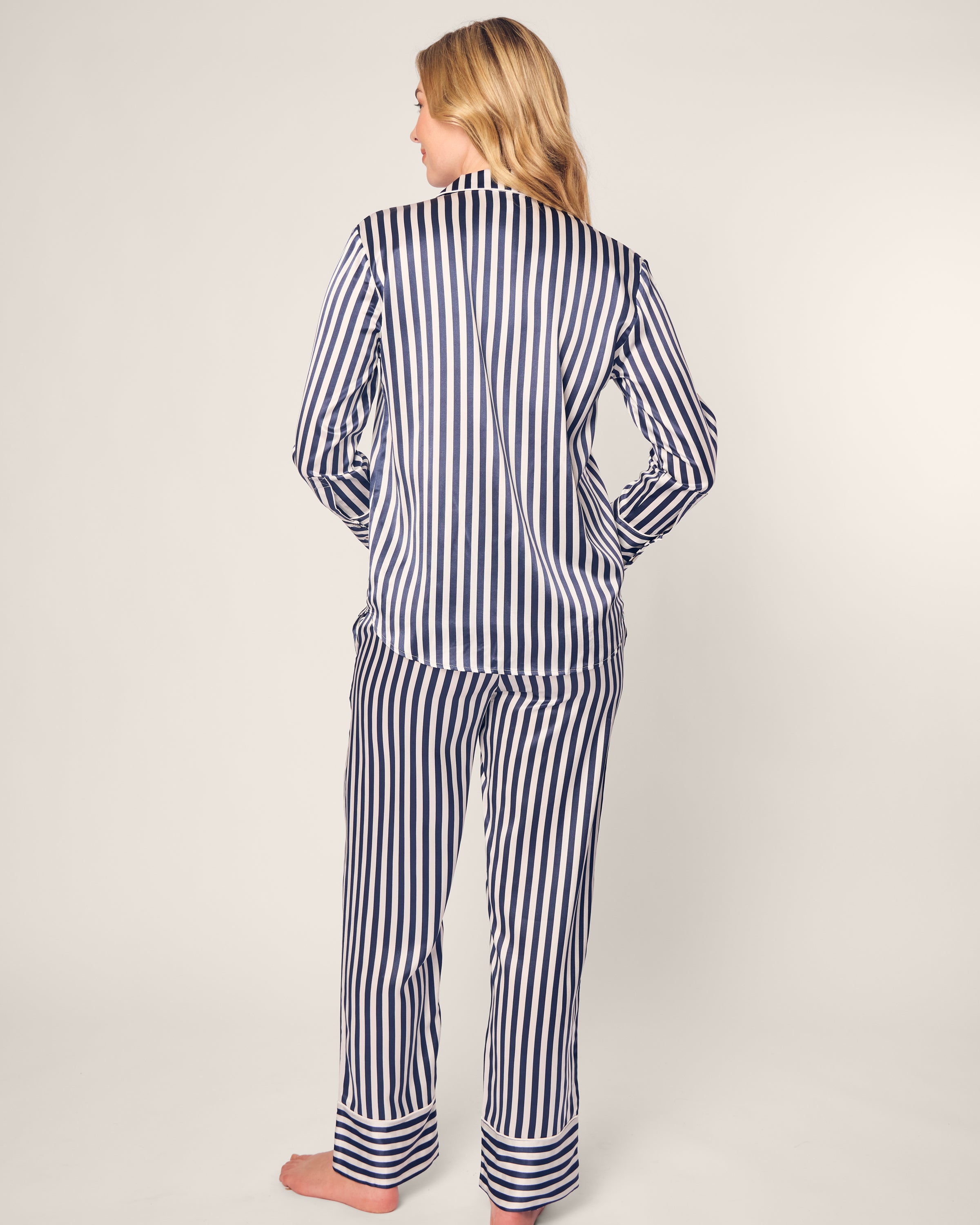 A person with long, blonde hair is wearing a Petite Plume Womens Silk Pajama Set in Navy Bengal Stripe, showcasing vertical blue and white stripes. They stand barefoot, facing away against a simple light background.