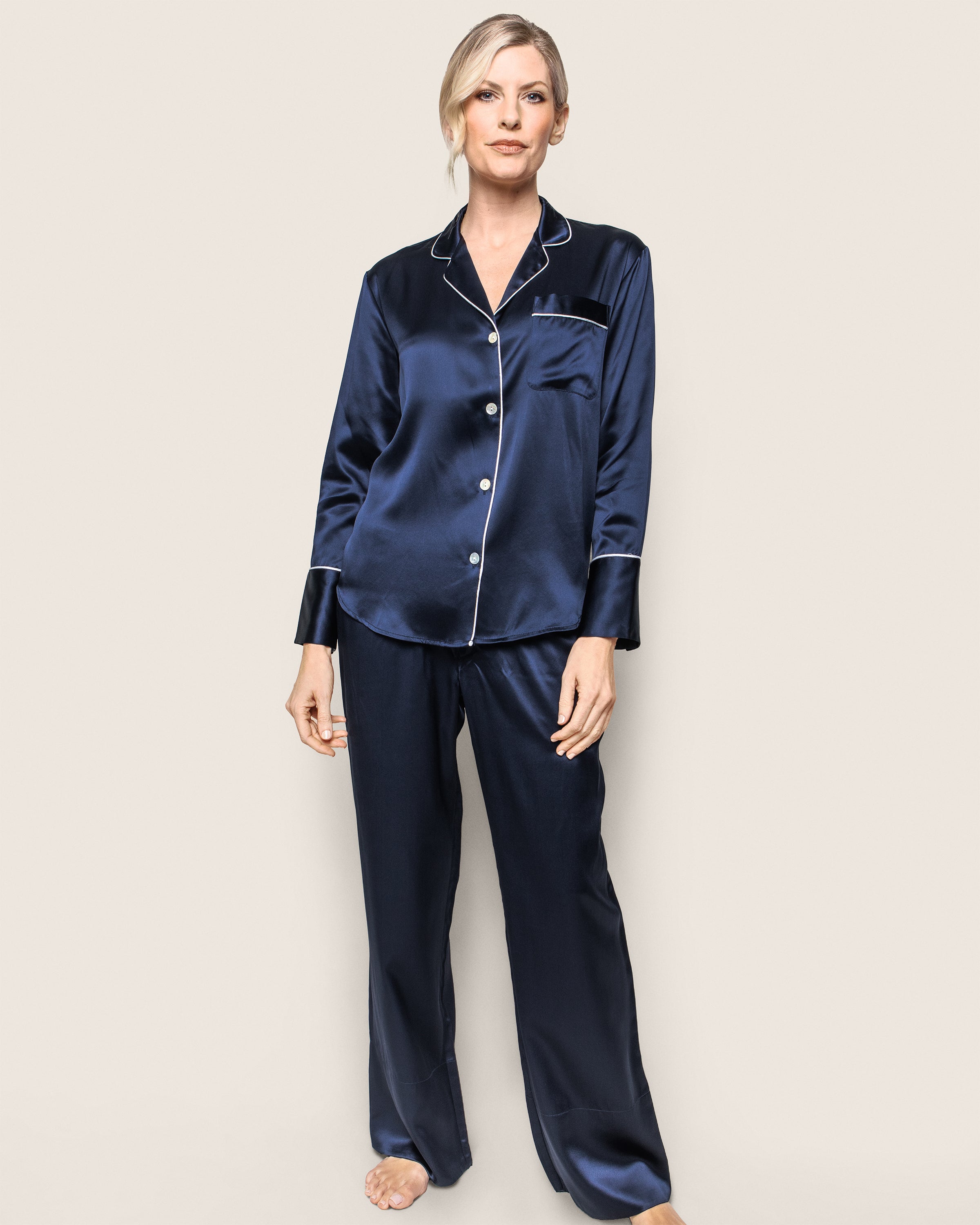 A person stands in Petite Plumes Womens Silk Pajama Set in Navy, showcasing luxurious sleepwear with white piping and a classic design—featuring a long-sleeve button-up top with a pocket and matching pants—against a plain background.