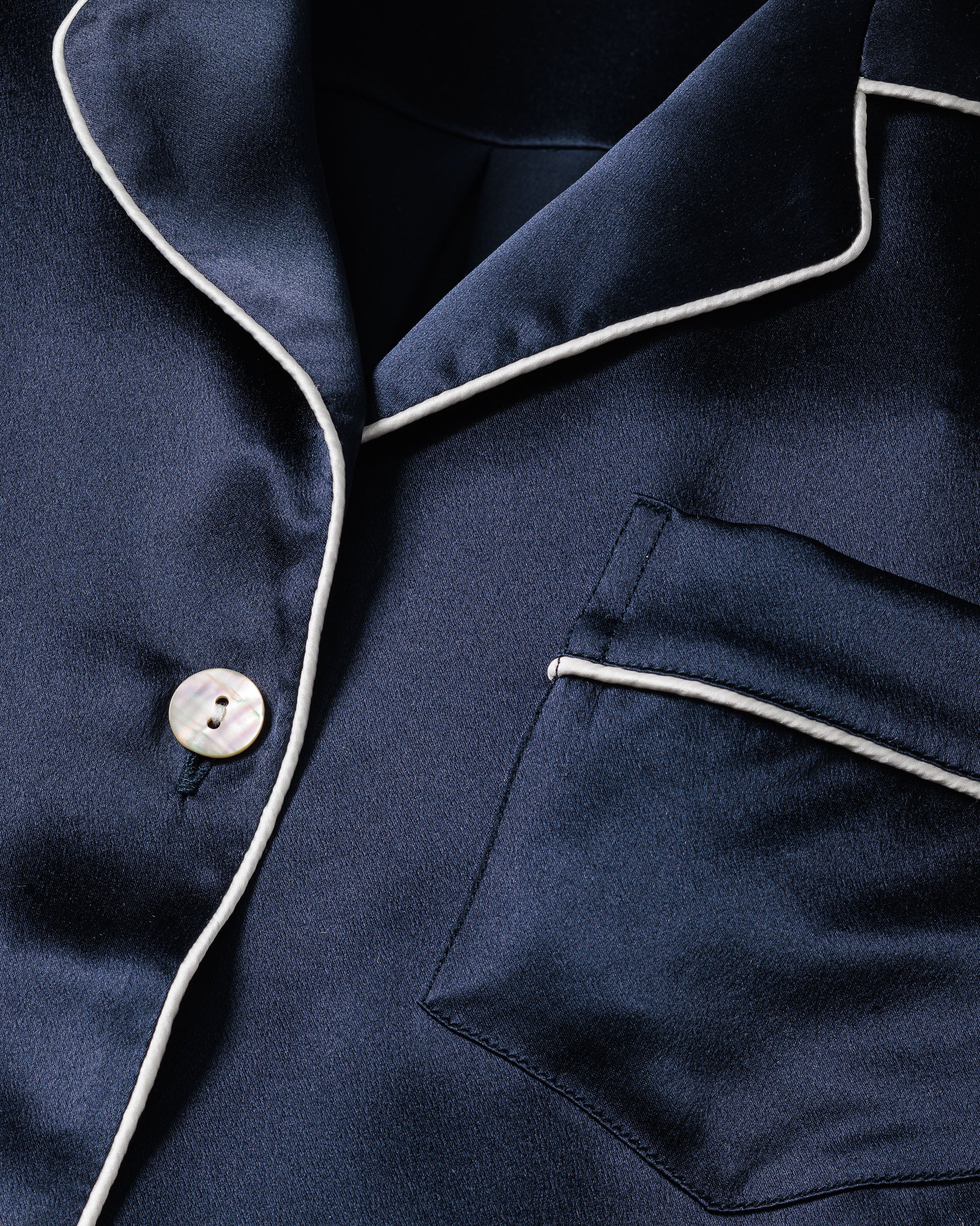 The Womens Silk Pajama Set in Navy by Petite Plume features a close-up of the navy blue silk top with white piping along the collar, pocket, and edges. This luxury sleepwear includes a front pocket and white button, offering a timeless silhouette and luxurious silk texture.