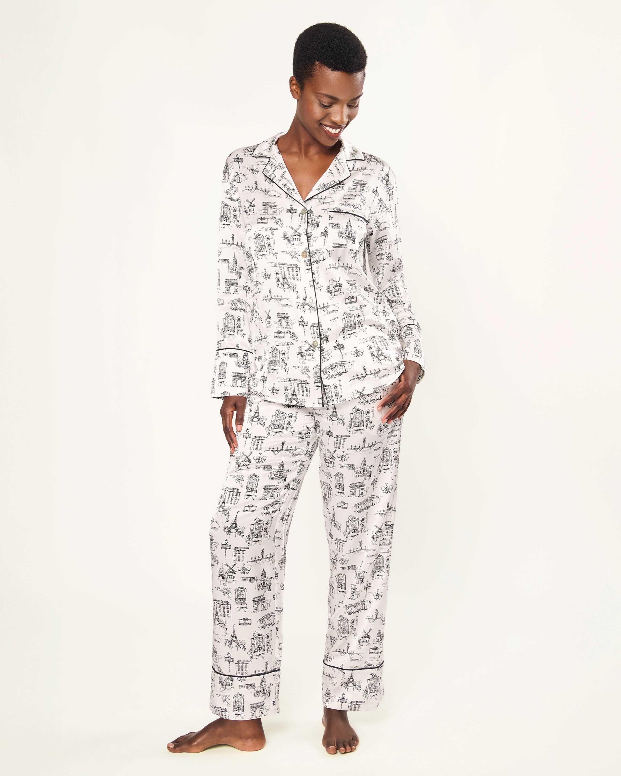 Women's Silk Pajama Set in Parisian Toile: white button-up with black piping, illustrated with buildings and trees. Includes long-sleeve top with chest pocket and matching long pants featuring an elastic waistband.