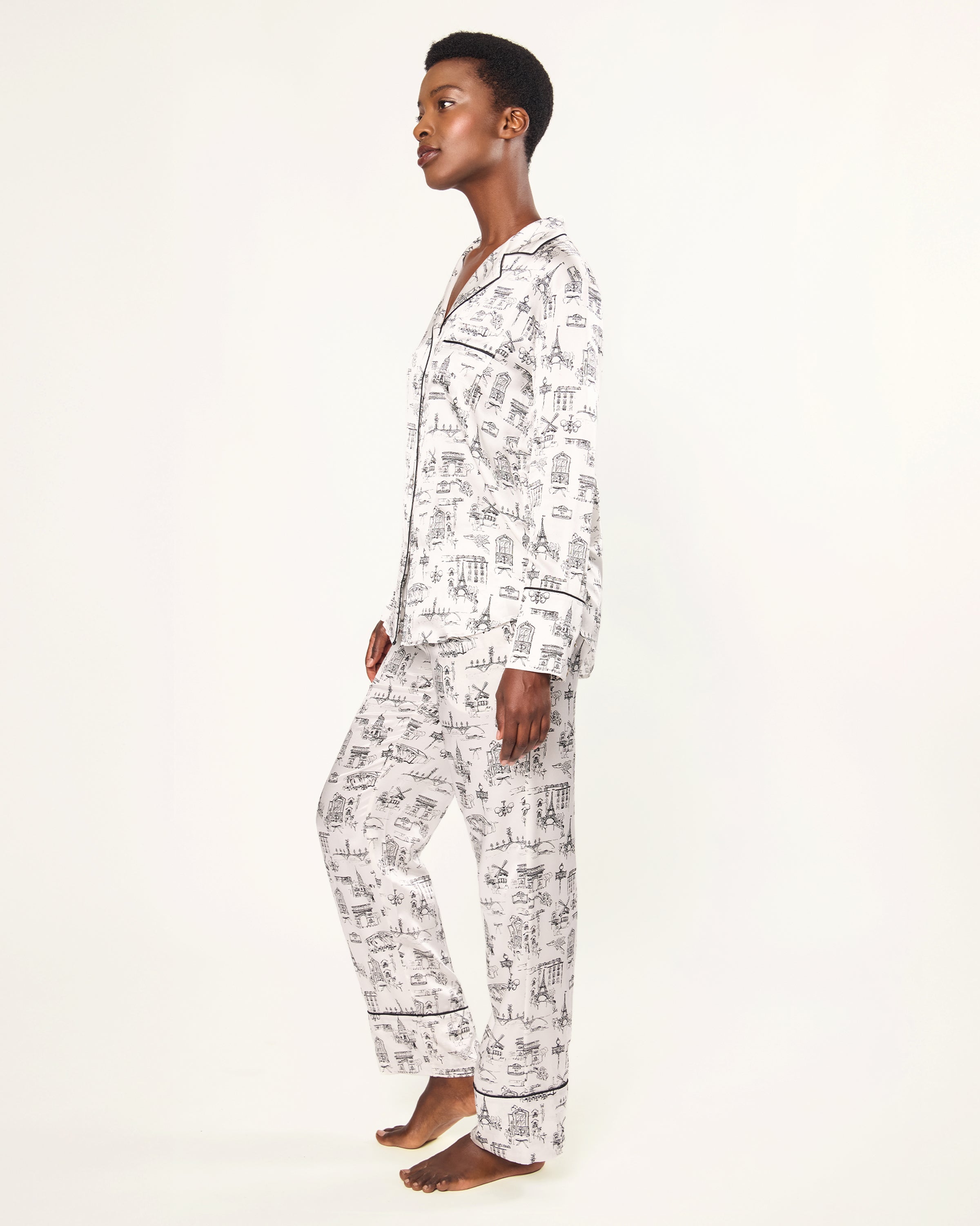 Women's Silk Pajama Set in Parisian Toile: white button-up with black piping, illustrated with buildings and trees. Includes long-sleeve top with chest pocket and matching long pants featuring an elastic waistband.