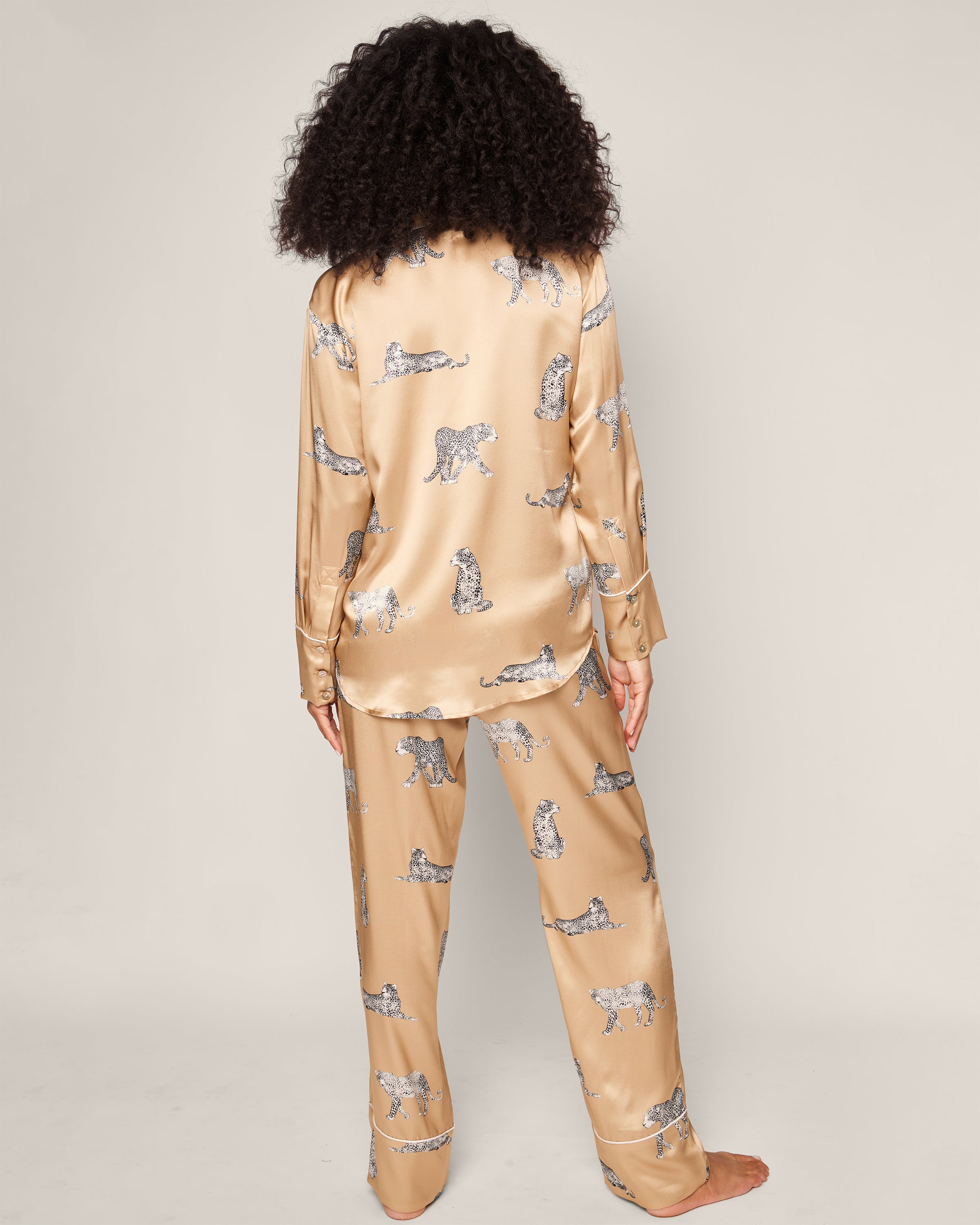 A person with curly hair stands barefoot, facing away, wearing Petite Plumes Womens Silk Pajama Set in Panthère Champagne. The luxury sleepwear is long-sleeved and full-length, made from Mulberry silk. The background is plain and neutral.