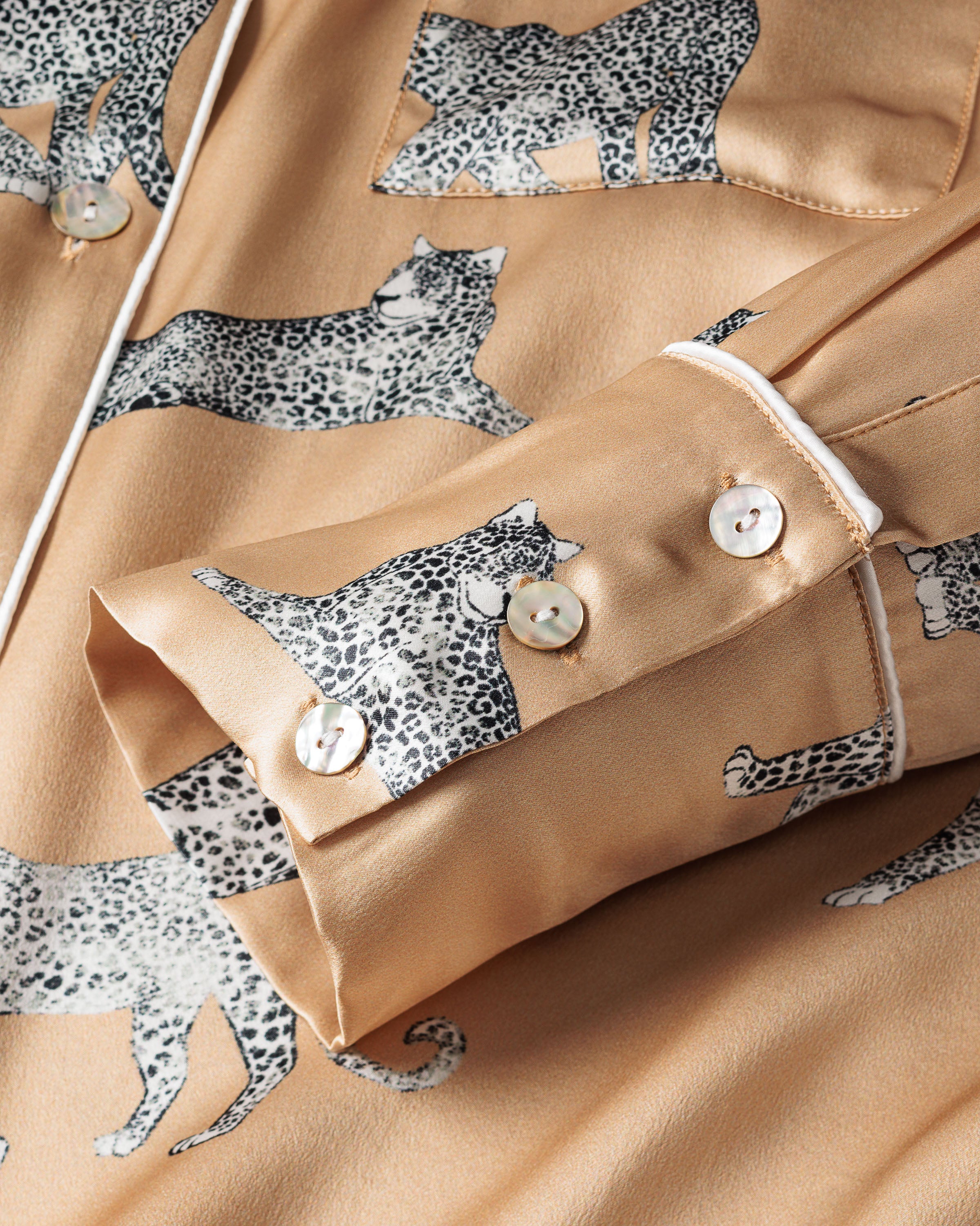A close-up of the Womens Silk Pajama Set in Panthère Champagne by Petite Plume shows a beige fabric with a black and white panther motif, exuding elegance. It features visible buttons and refined white piping along the cuff edge.