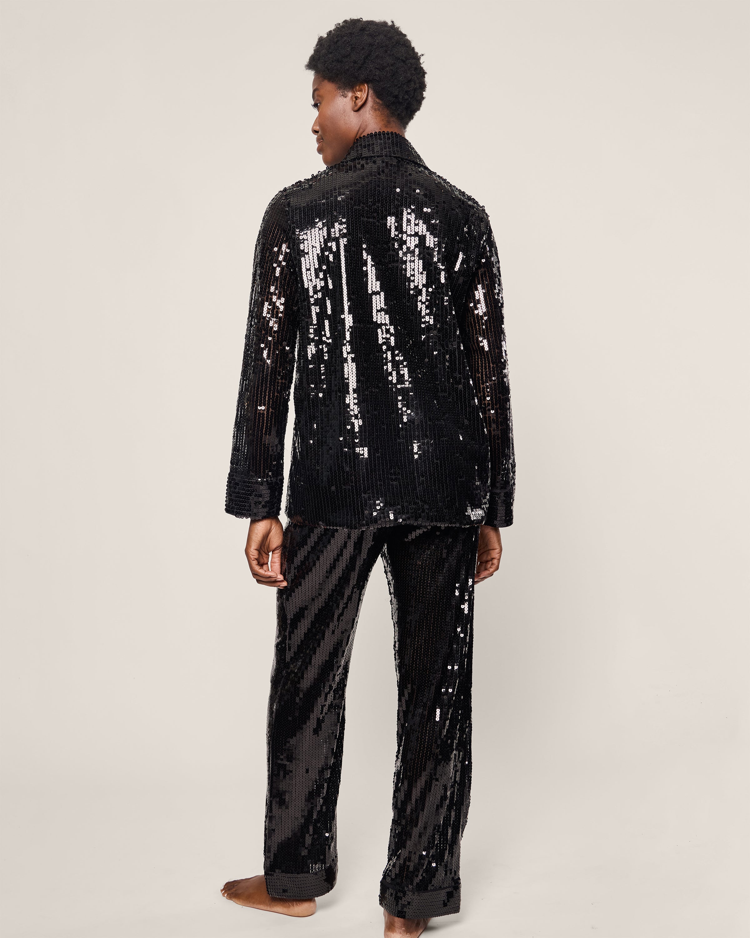 A person in Petite Plumes Womens Pajama Set in Black Sequins stands barefoot against a plain backdrop, slightly turning away from the camera. The reflective long sleeves and full-length pants blend luxury sleepwear with captivating style.
