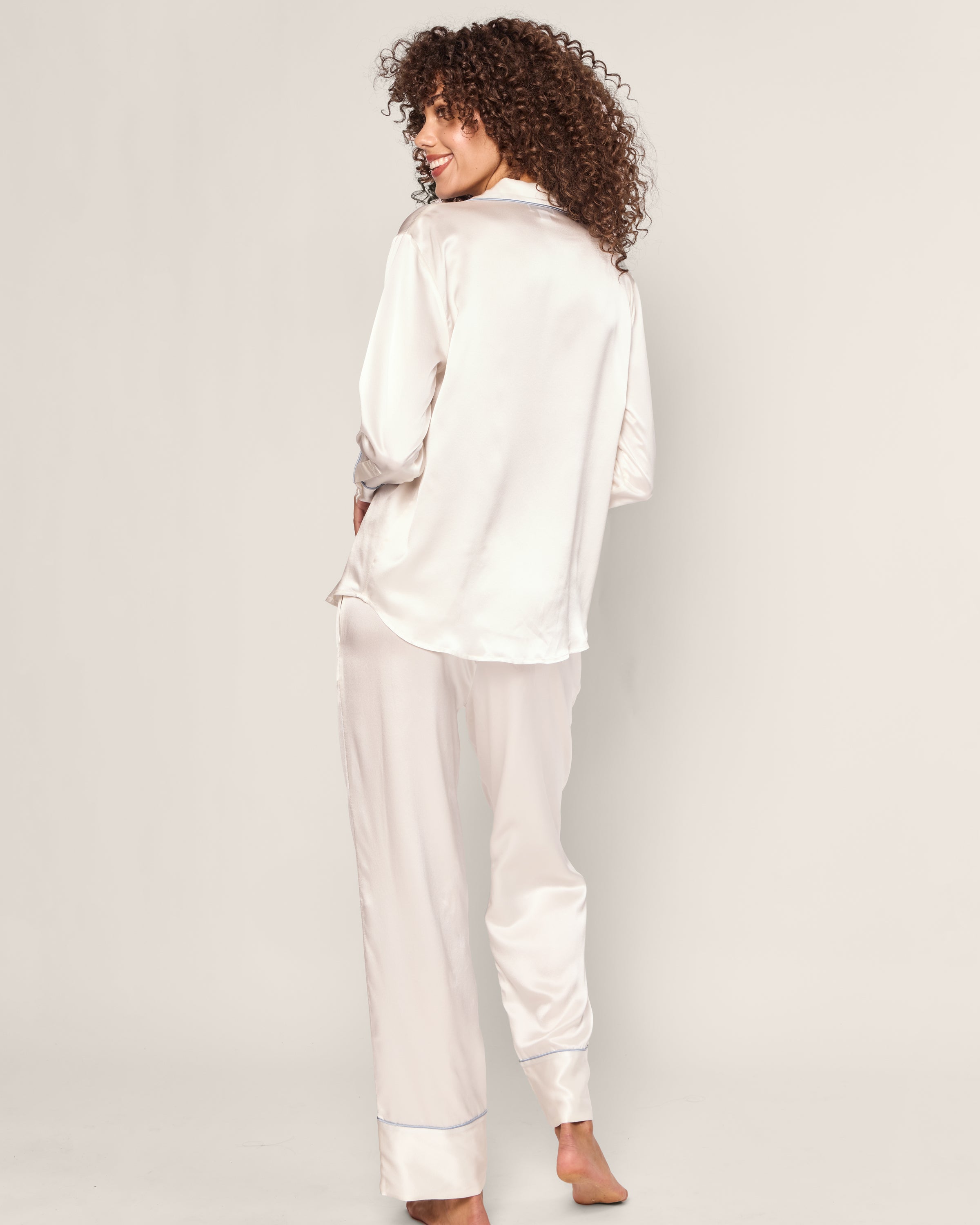 A person with curly hair faces away, wearing a Petite Plume Womens Silk Pajama Set in white with periwinkle piping. The set includes a long-sleeved top and loose pants made of luxurious mulberry silk, against a plain light-colored background.