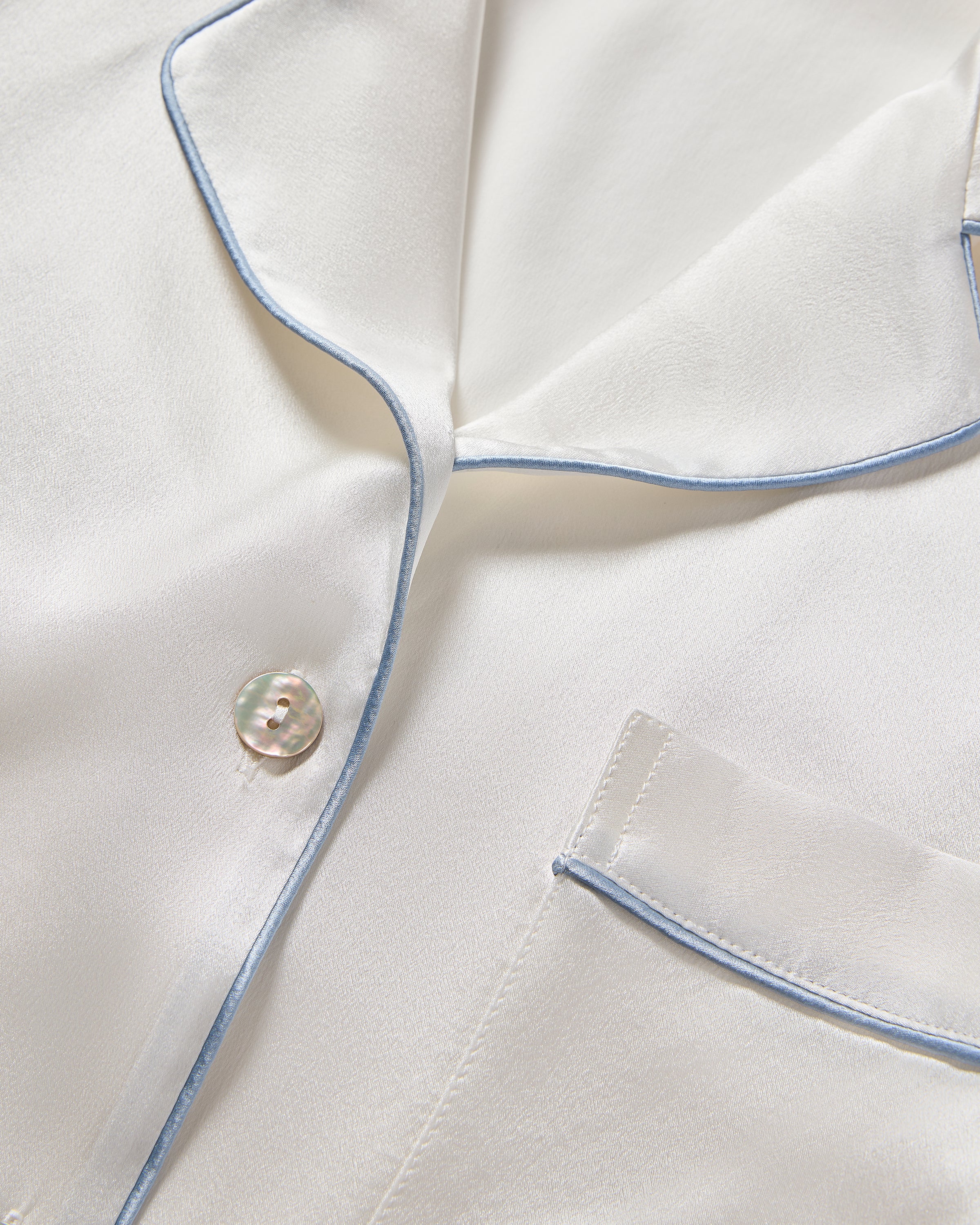 Close-up of Petite Plumes Womens Silk Pajama Set in white mulberry silk with periwinkle piping along the collar and chest pocket. This elegant, timeless piece features a visible button and doubles as a bridal keepsake.