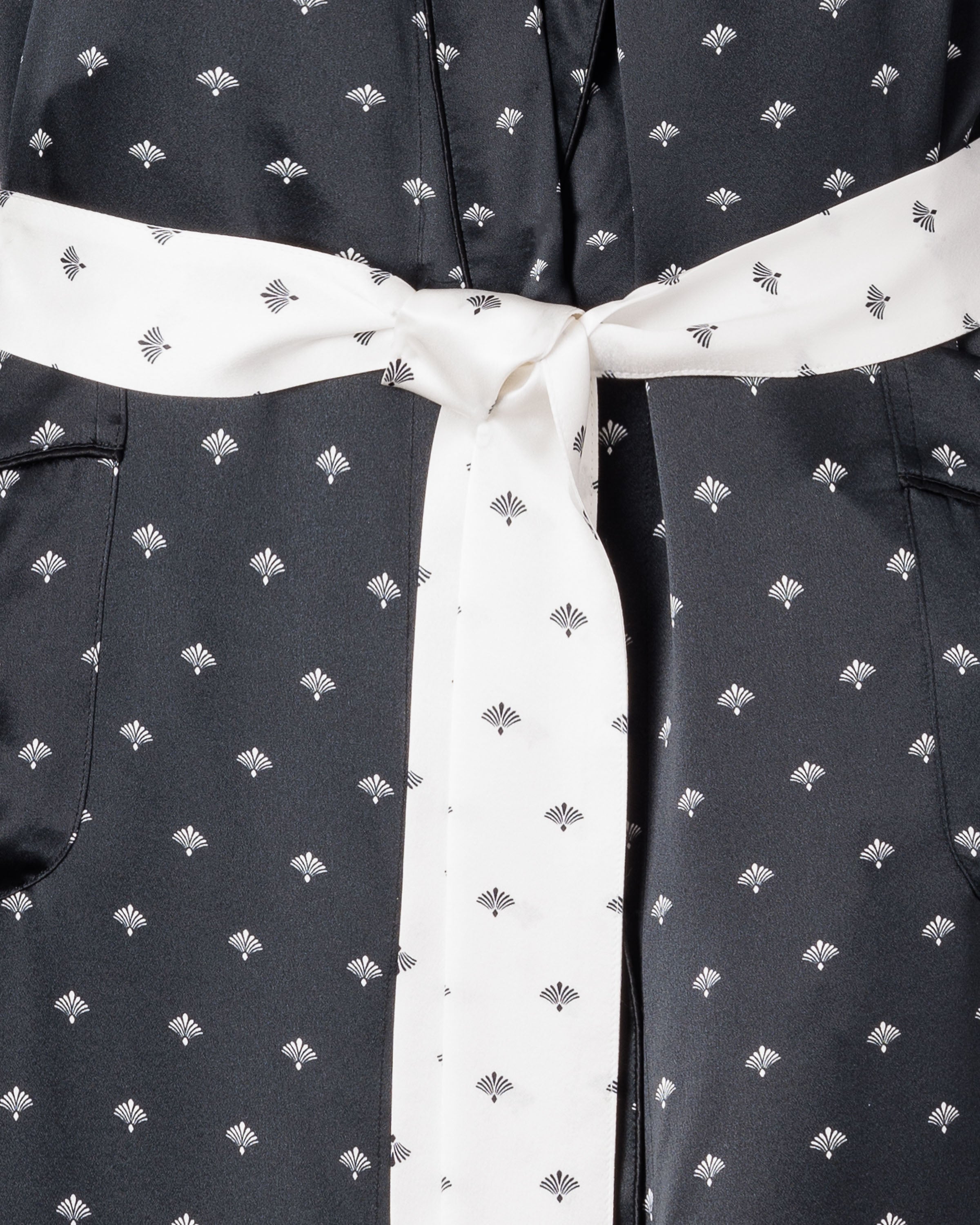 The Womens Silk Long Robe in Black Art Nouveau by Petite Plume features a close-up black fabric with small white geometric patterns reminiscent of an Art Deco fan motif and a contrasting white sash tied in a bow at the center, reminiscent of Truman Capotes style.
