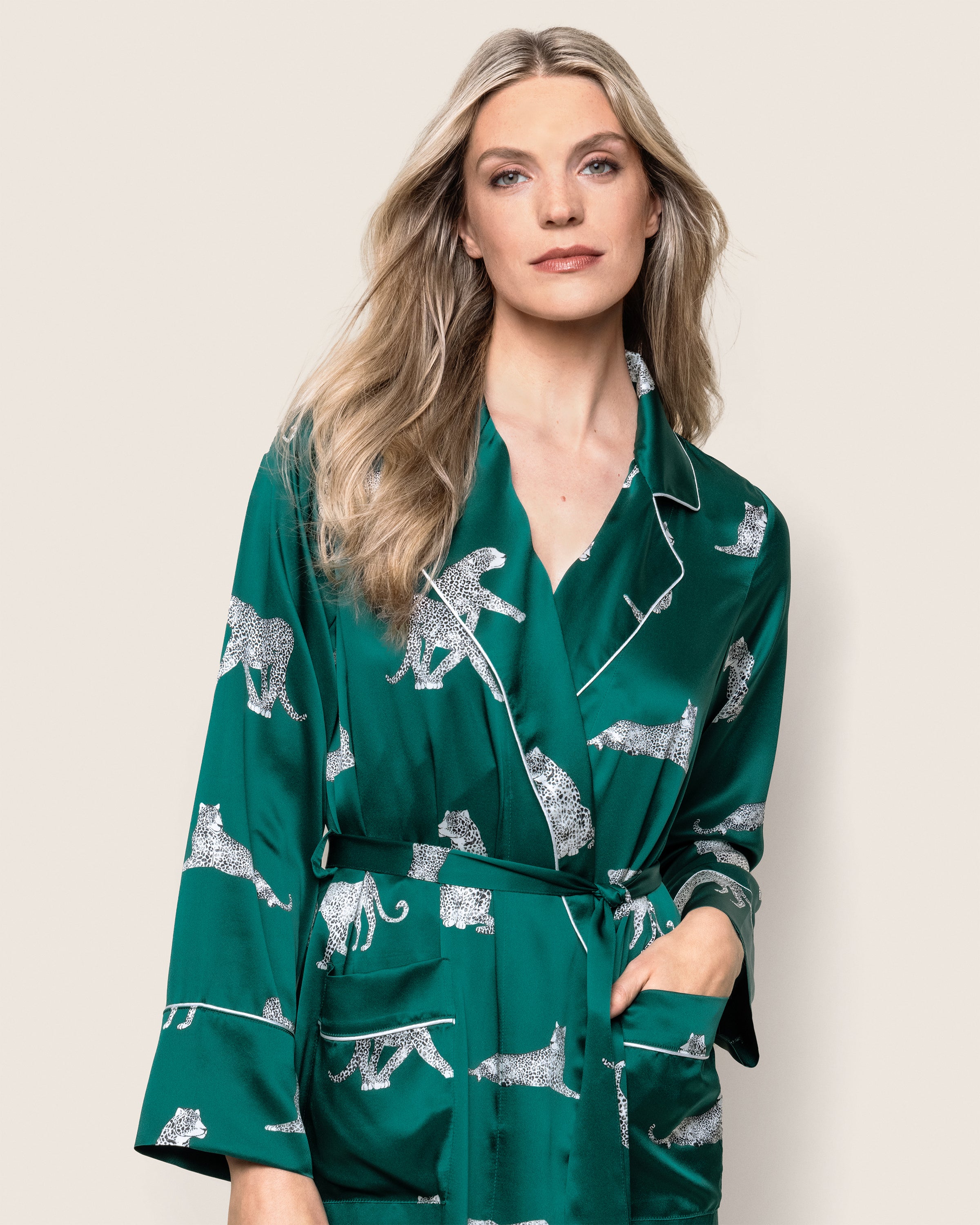 A person with long, light hair wearing Petite Plumes Womens Silk Long Robe in Panthère de Luxe, a green mulberry silk robe adorned with white leopard illustrations, stands against a plain background, facing the camera.