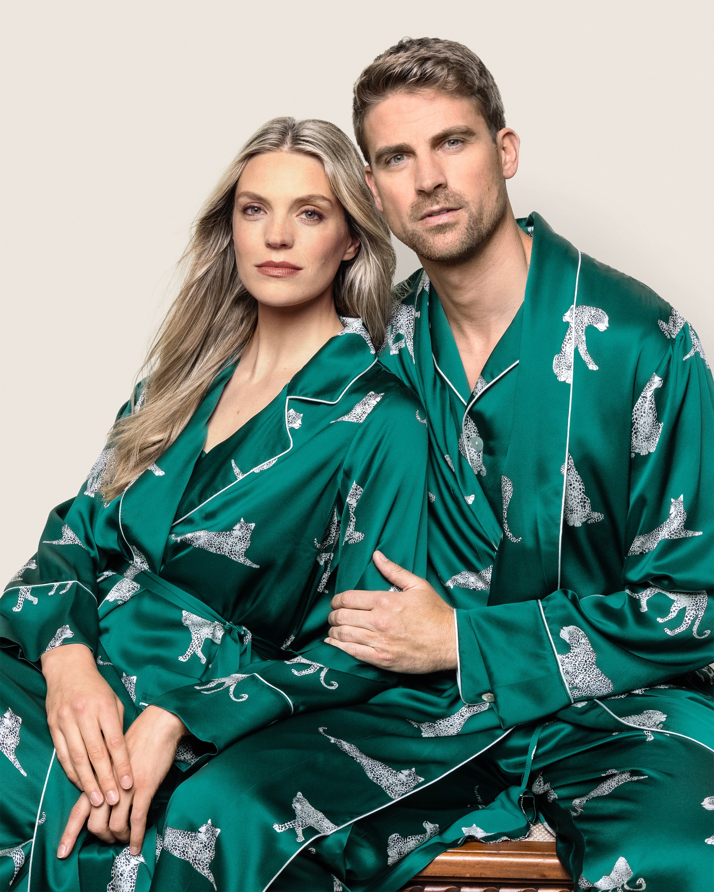 A couple, the woman with long blonde hair in Petite Plumes Womens Silk Long Robe in Panthère de Luxe, and the man with short brown hair, sit closely. His arm around her shoulders showcases their chic mulberry silk sleepwear against a plain background.