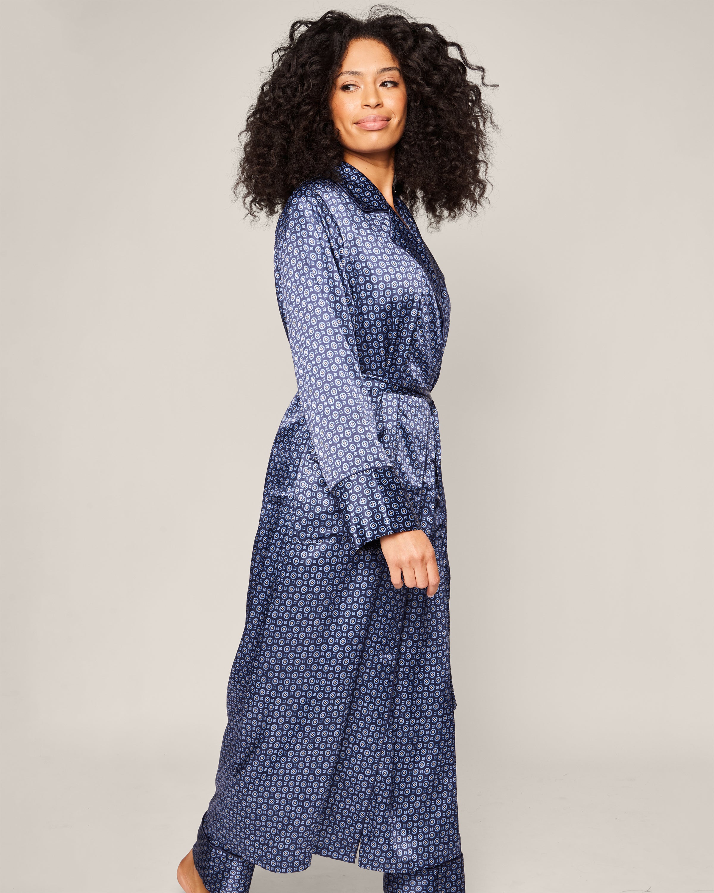 A curly-haired person wears Petite Plumes Womens Silk Robe in Midnight Foulard with matching pants, walking left against a plain background. They display a content expression while styling a neck scarf effortlessly.