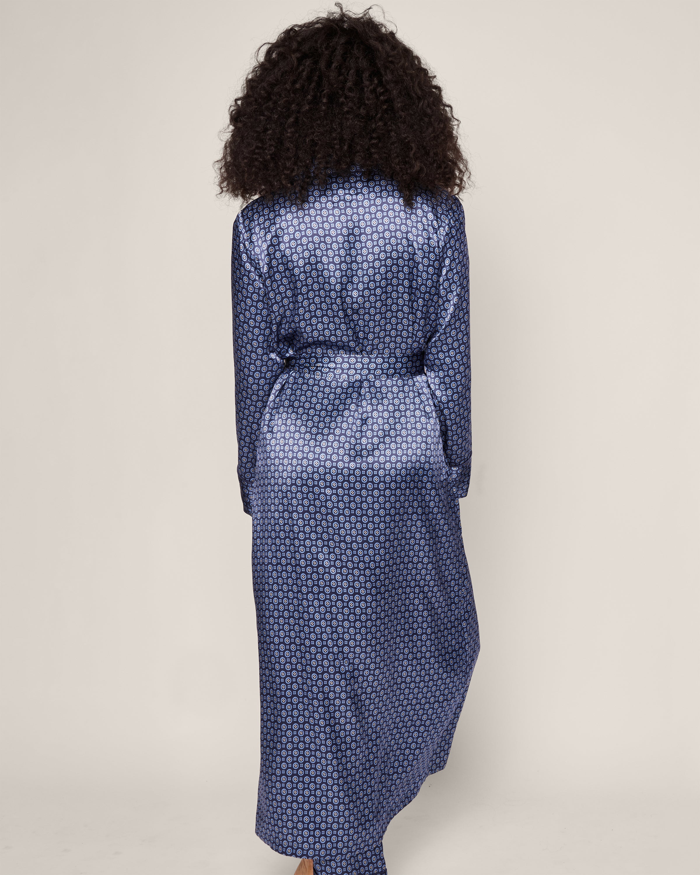 A person with curly hair stands with their back to the camera against a plain background, wearing Petite Plumes Womens Silk Robe in Midnight Foulard, complemented by a mulberry silk neck scarf.