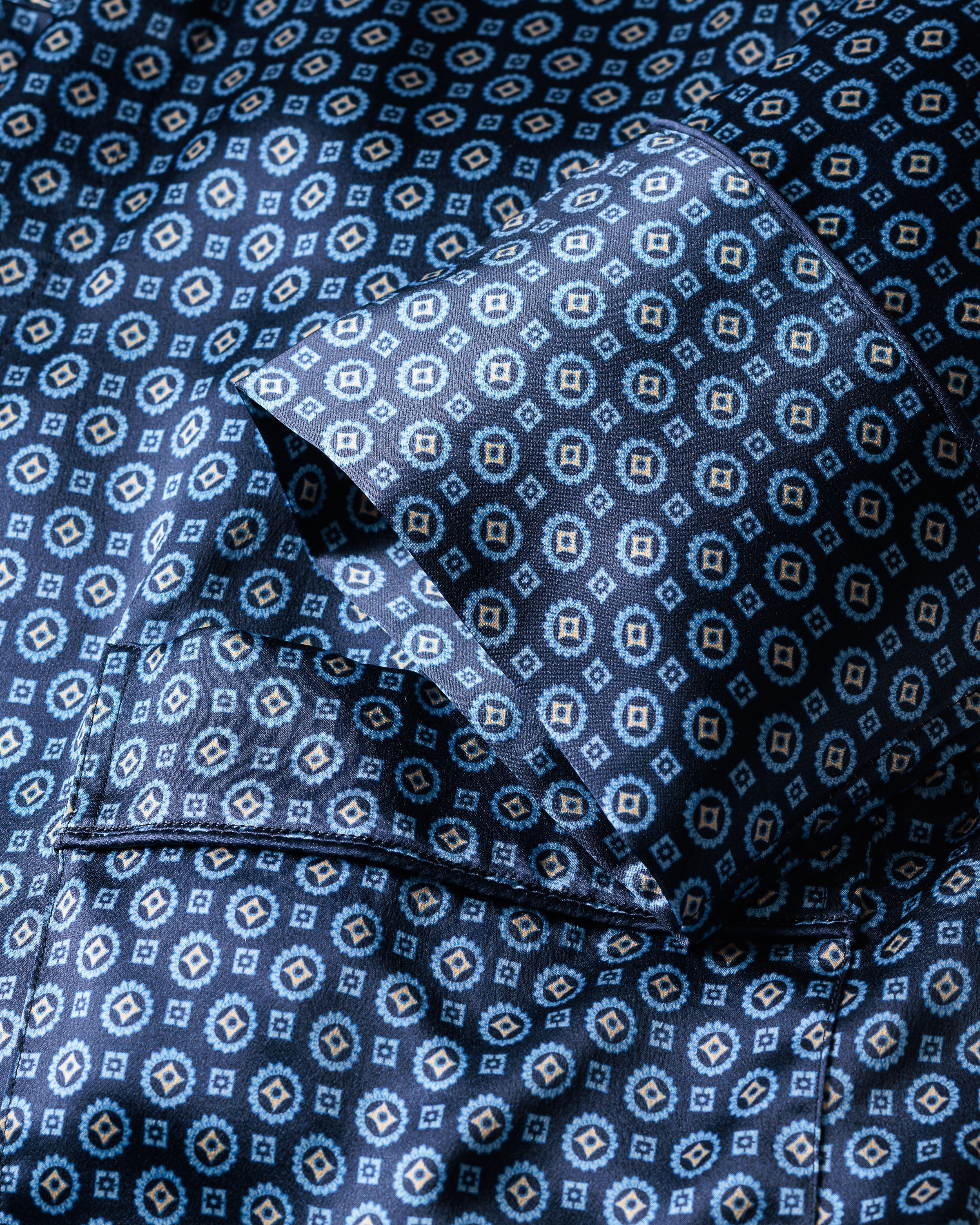 A close-up of Petite Plumes Womens Silk Robe in Midnight Foulard shows its blue fabric with a white and beige geometric pattern. Made from Mulberry silk, it has a crisp texture and folded sleeves, offering a modern design ideal for sophisticated sleepwear or accessorizing with an elegant neck scarf.