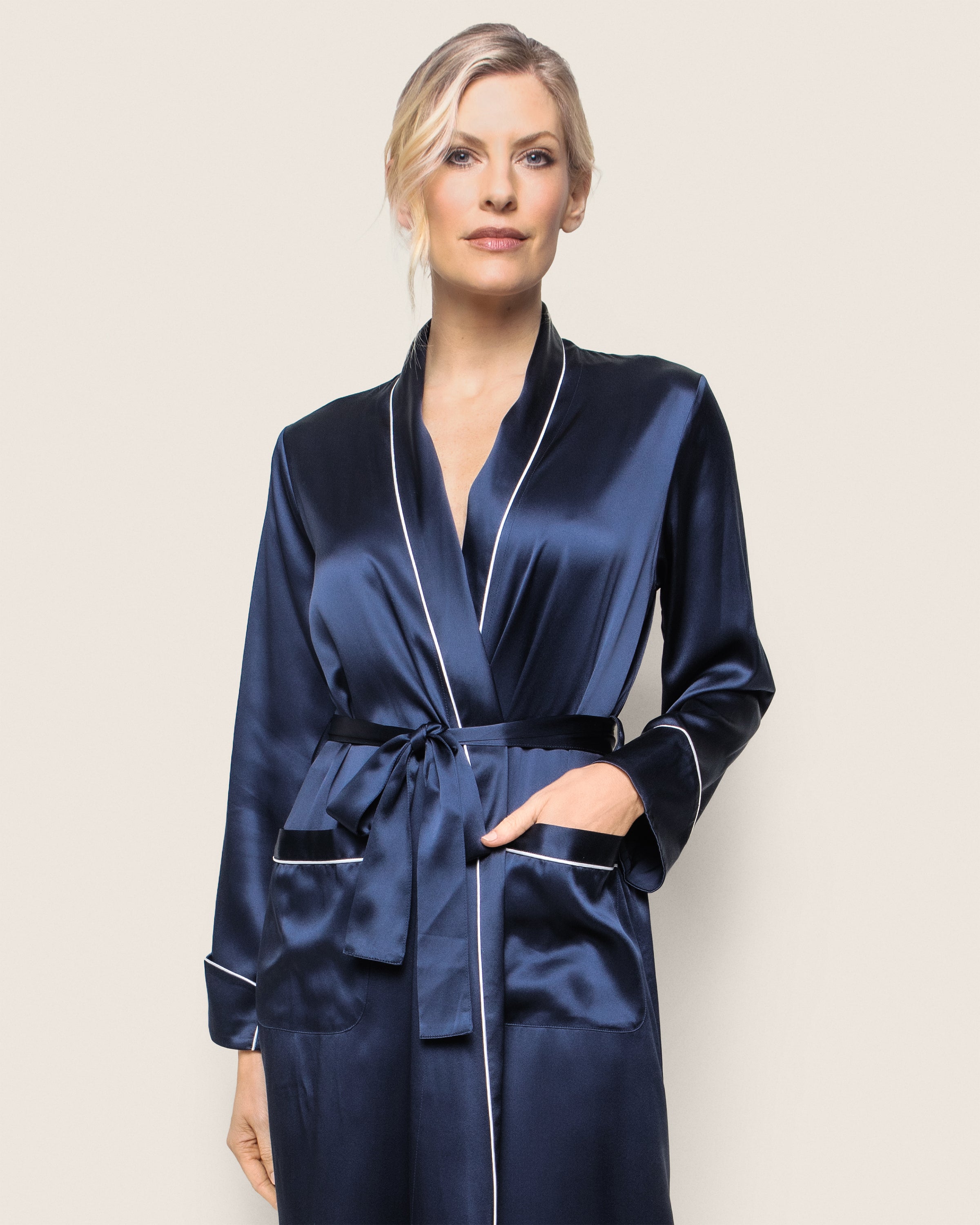A person models the Womens Silk Long Robe in Navy by Petite Plume. This luxurious robe features white piping, is crafted from 22mm silk, and tied with a matching belt. The individual stands against a light background with one hand in a pocket, epitomizing elegant luxury sleepwear.