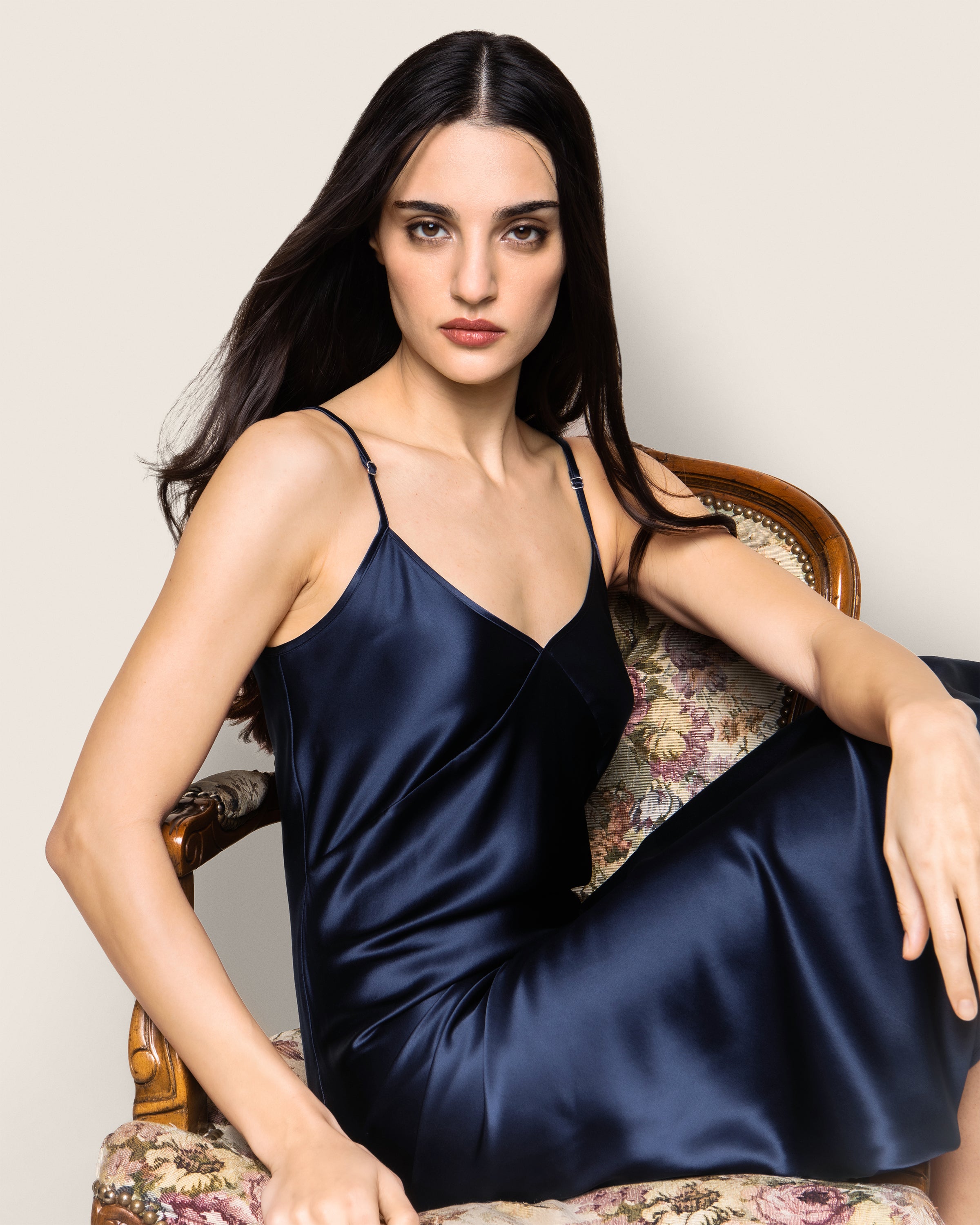 A woman with long dark hair sits on a floral chair wearing the Petite Plume Womens Silk Cosette Nightgown in Navy. She gazes at the camera neutrally, her right arm resting on the armrest. The background is plain and neutral.