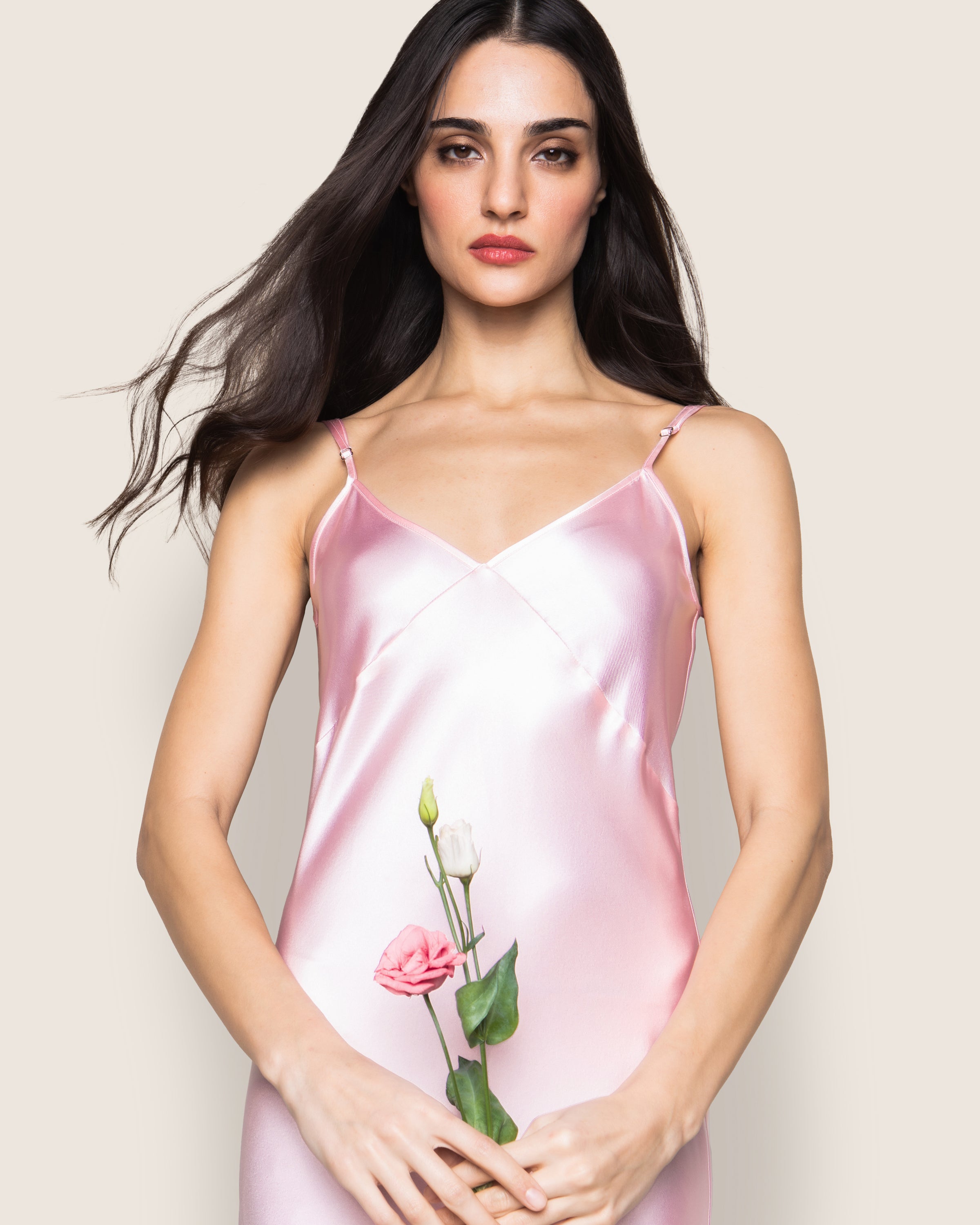 A woman with long dark hair, wearing the Petite Plume Womens Silk Cosette Nightgown in Pink, holds a small bouquet of pink and white flowers against a plain, light-colored background, evoking a minimalist fantasy.