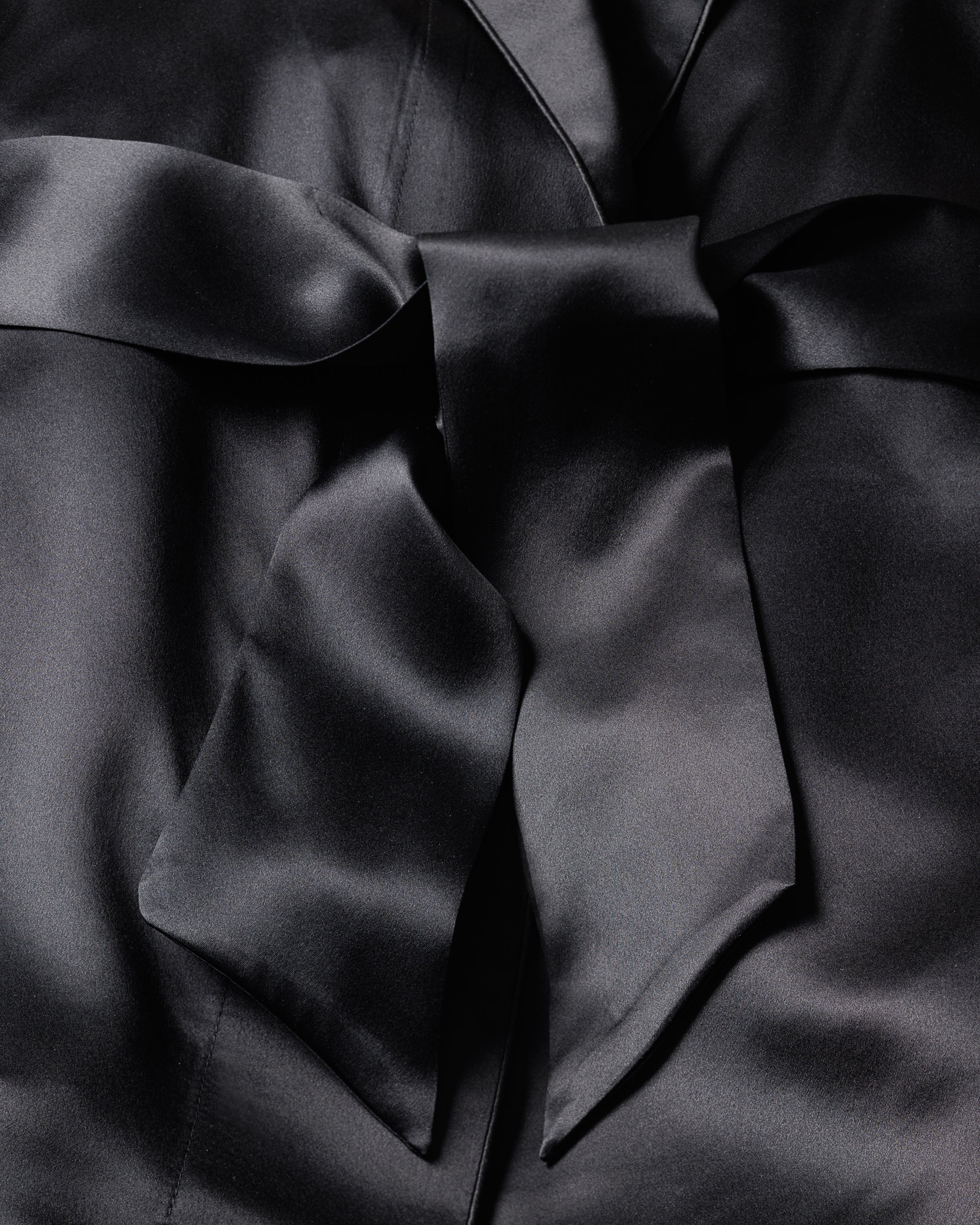 The Womens Silk Short Robe in Black by Petite Plume features smooth, glossy black satin reminiscent of luxe sleepwear. Its delicate folds and creases highlight an elegant, hypoallergenic appearance tied into a chic bow.