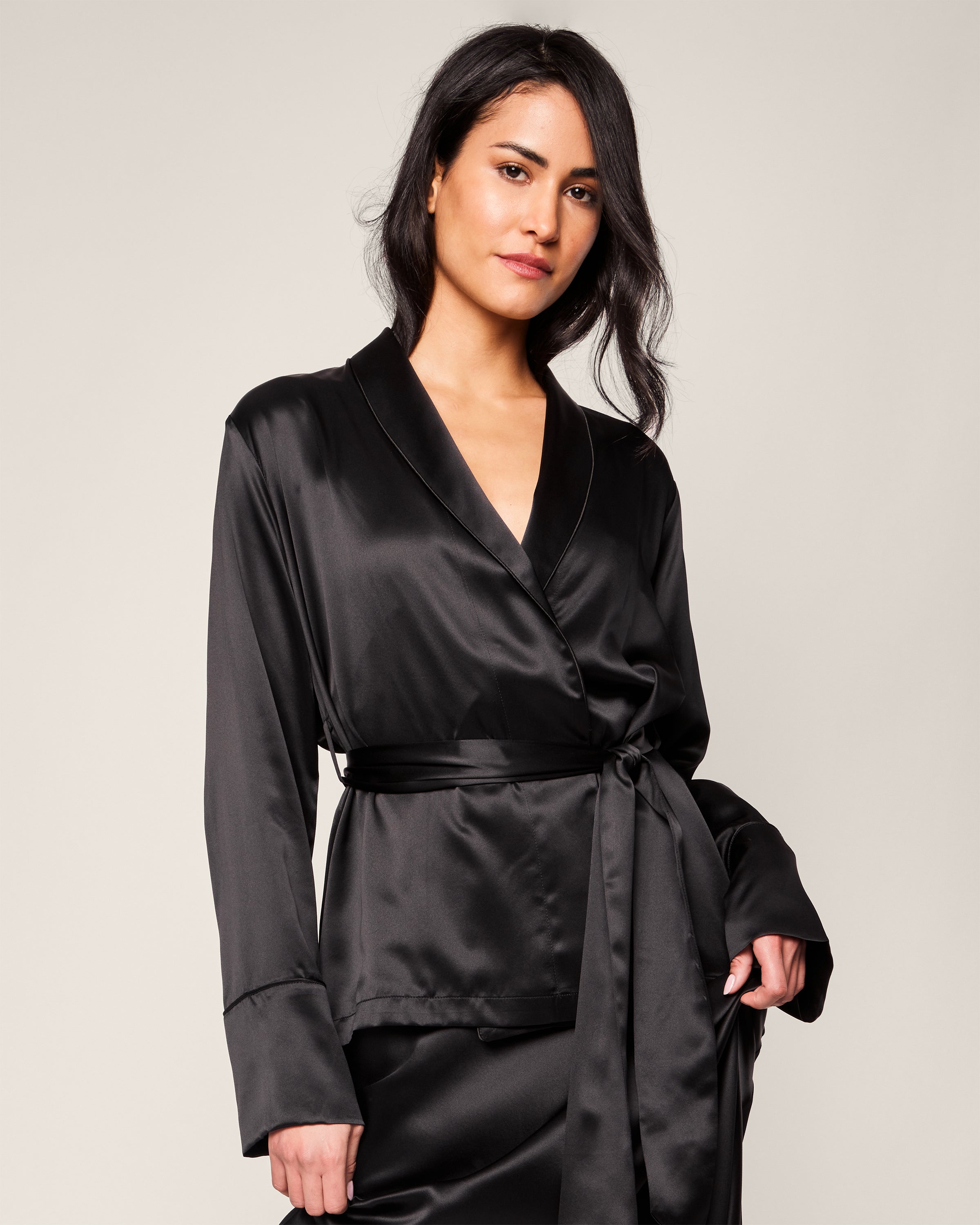 A woman poses against a neutral background in a black Womens Silk Short Robe by Petite Plume. Her hair flows in loose waves, exuding confidence. The robes sleek tie-waist design offers an elegant look reminiscent of luxurious sleepwear.